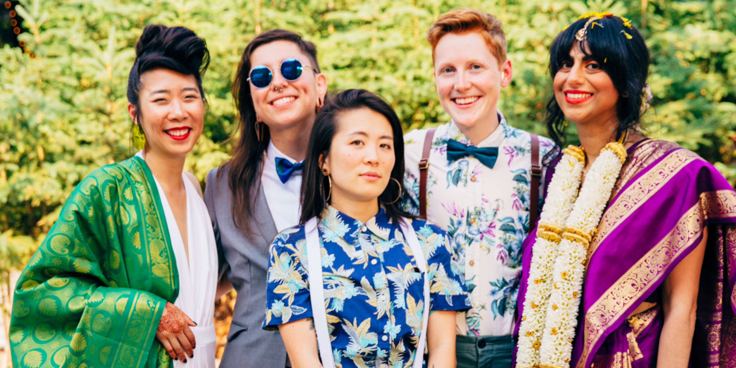 A group of five people standing outdoors in front of greenery. They are smiling and dressed in a variety of colorful outfits, including floral shirts, a gray suit, and traditional attire with garlands. One person is wearing sunglasses.