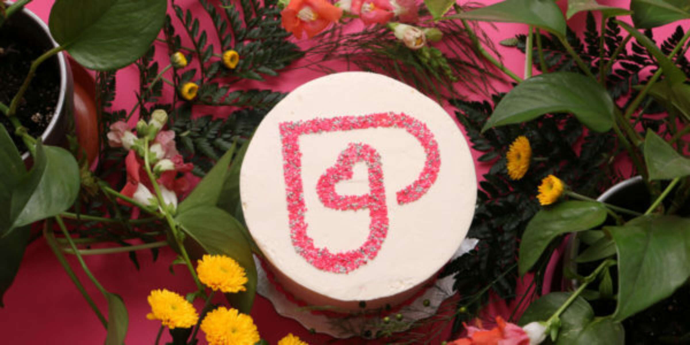 A round, white cake sits on a pink surface, surrounded by a variety of flowers and greenery. The cake is decorated with a red sugar outline of an abstract heart shape on top. Yellow and orange blooms are prominent among the surrounding foliage.