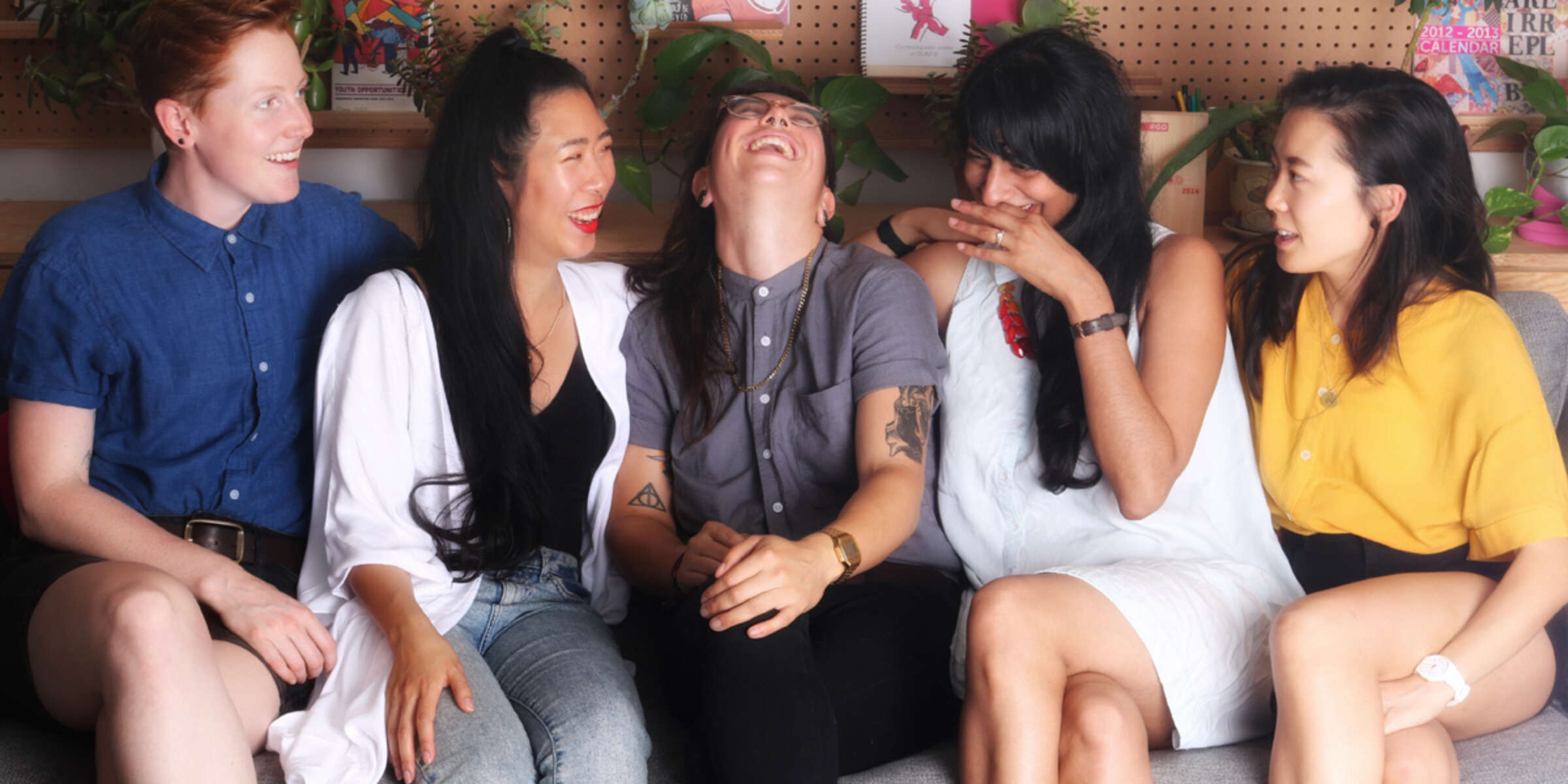 A group of five women sitting on a couch, smiling and laughing together. They are dressed in casual clothing and appear to be sharing a joyful moment. The background features shelves with books and plants.