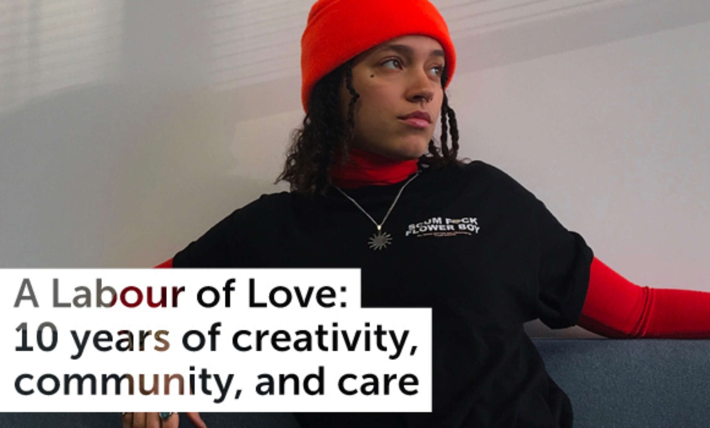 Person wearing an orange beanie, red long-sleeve shirt, and black t-shirt with a graphic design, sitting on a couch and looking to the side. Text overlay reads: "A Labour of Love: 10 years of creativity, community, and care.