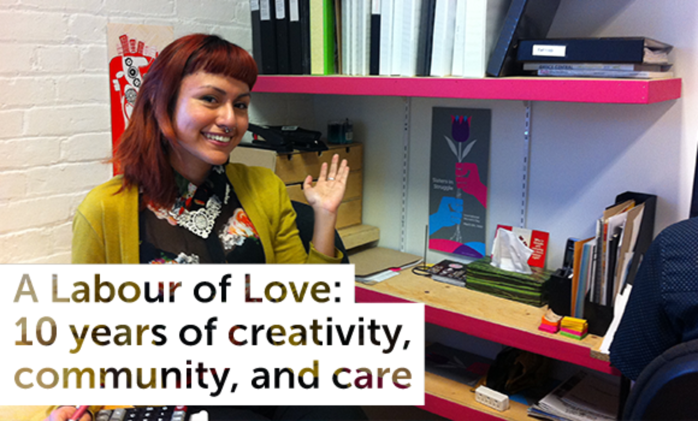 A person with reddish hair smiles and waves while sitting at a desk surrounded by books, folders, and office supplies. The overlay text reads, "A Labour of Love: 10 years of creativity, community, and care.