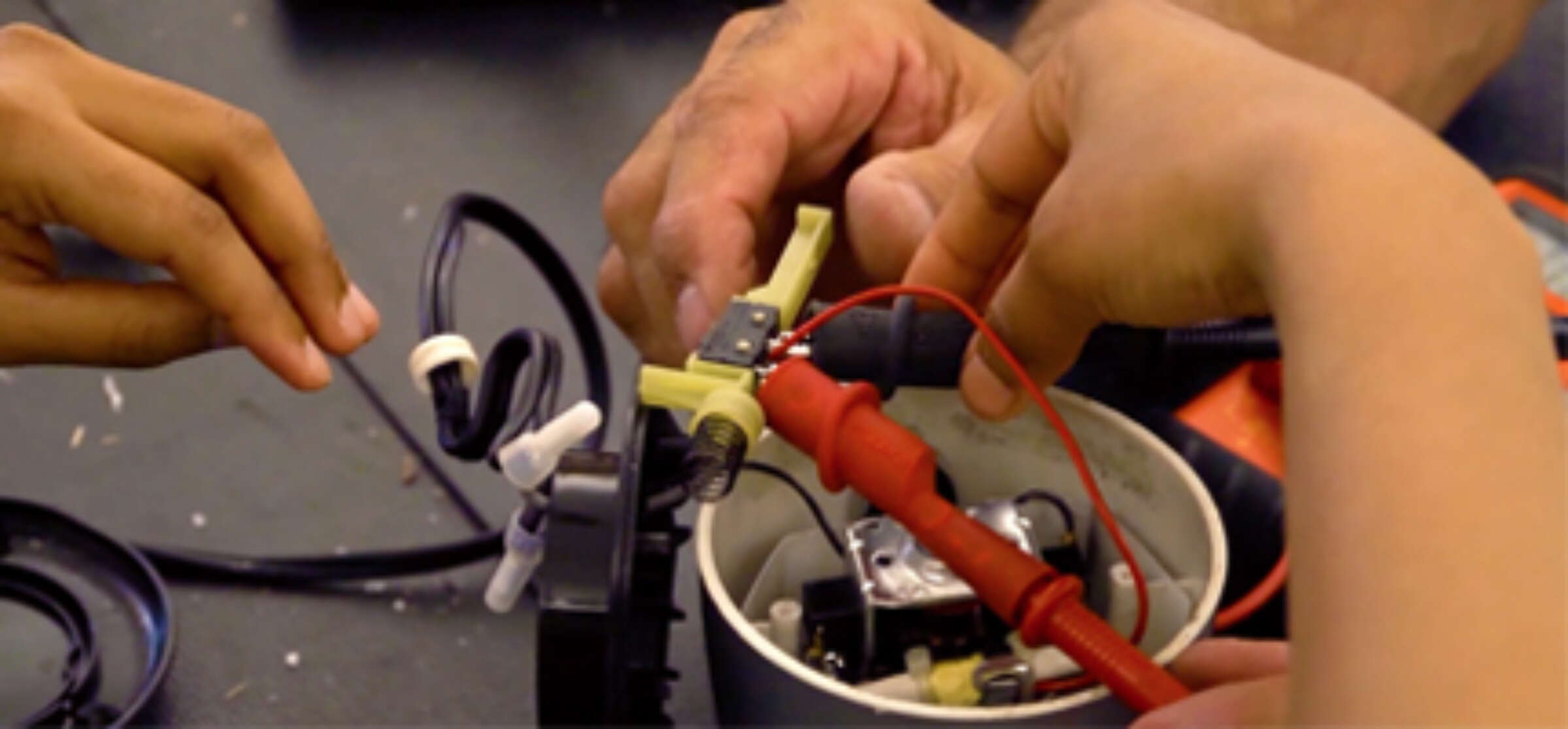 A close-up of a hands-on electronics project. Several hands are working together to assemble components including wires, a circuit board, and a switch, inside a small container. Tools and wires can be seen on the table around the project.