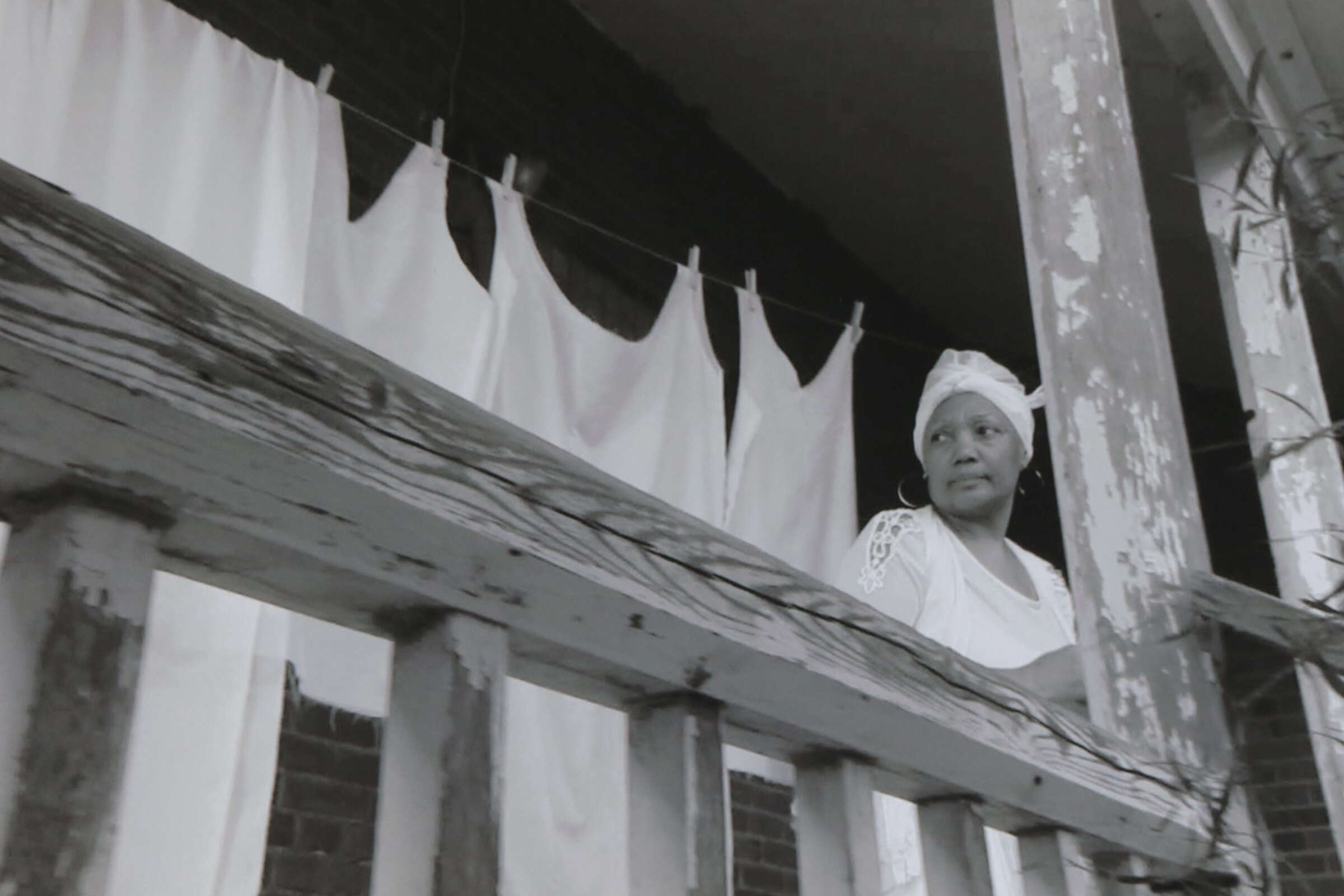 A black-and-white photo of a woman standing on a wooden porch. She looks pensively to the side, wearing a headscarf and a white top. Behind her, white sheets hang on a clothesline. The porch railings show signs of wear and age.