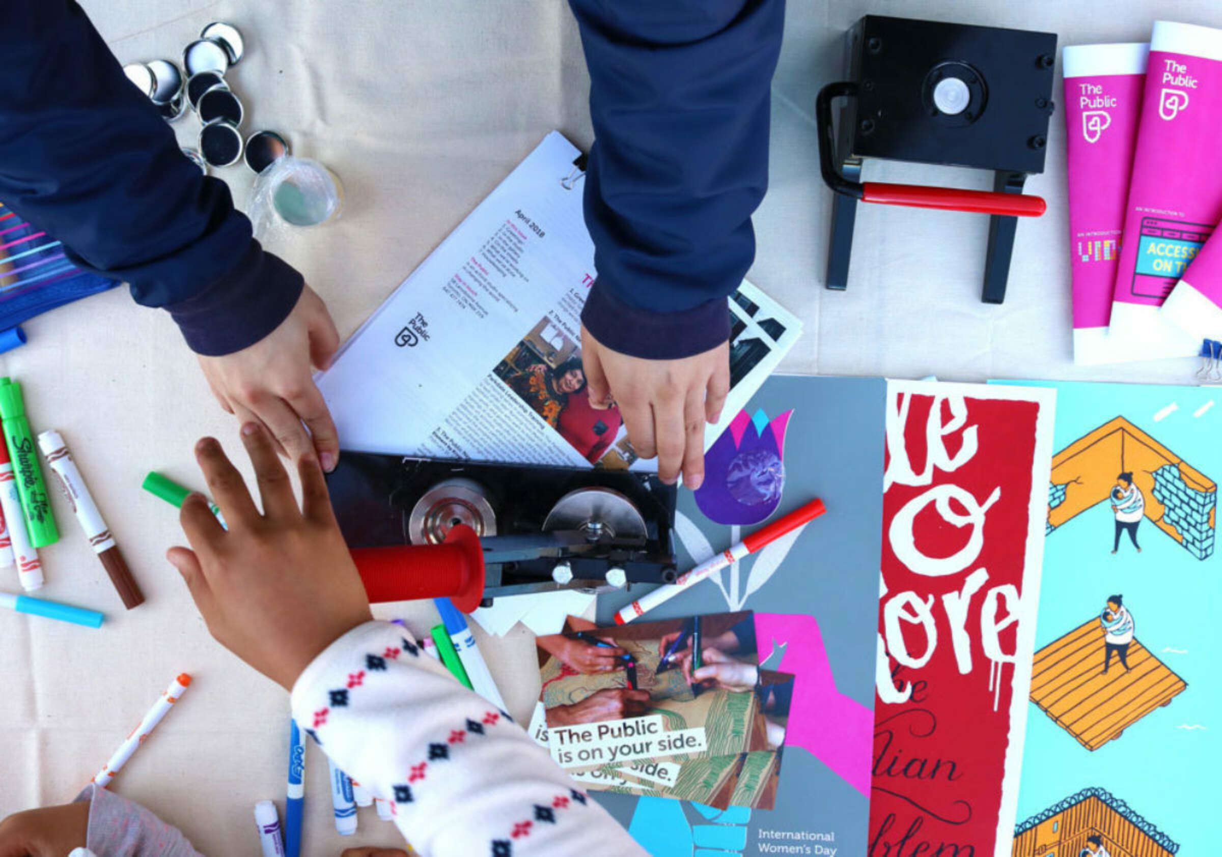 Three people work on an arts and crafts project on a table, with various supplies scattered around, including markers, magazines, and a book binding press. Their hands are engaged with the materials, and some colorful artwork is visible on the table.