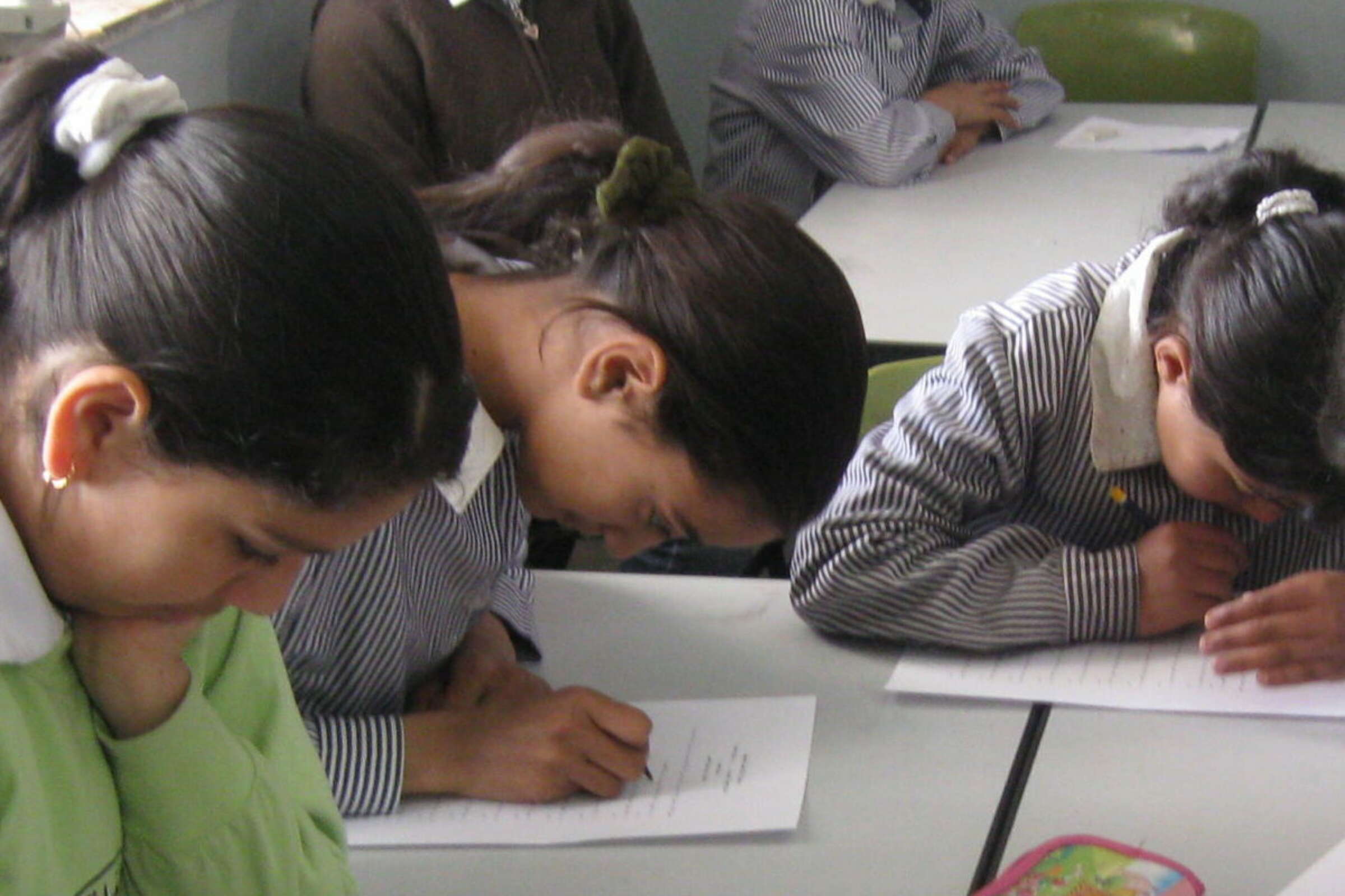 Three children with dark hair are sitting at a white table, leaning over their papers and writing with concentration. They are wearing striped uniforms. The background shows another student who is not in focus, seated at the same table.