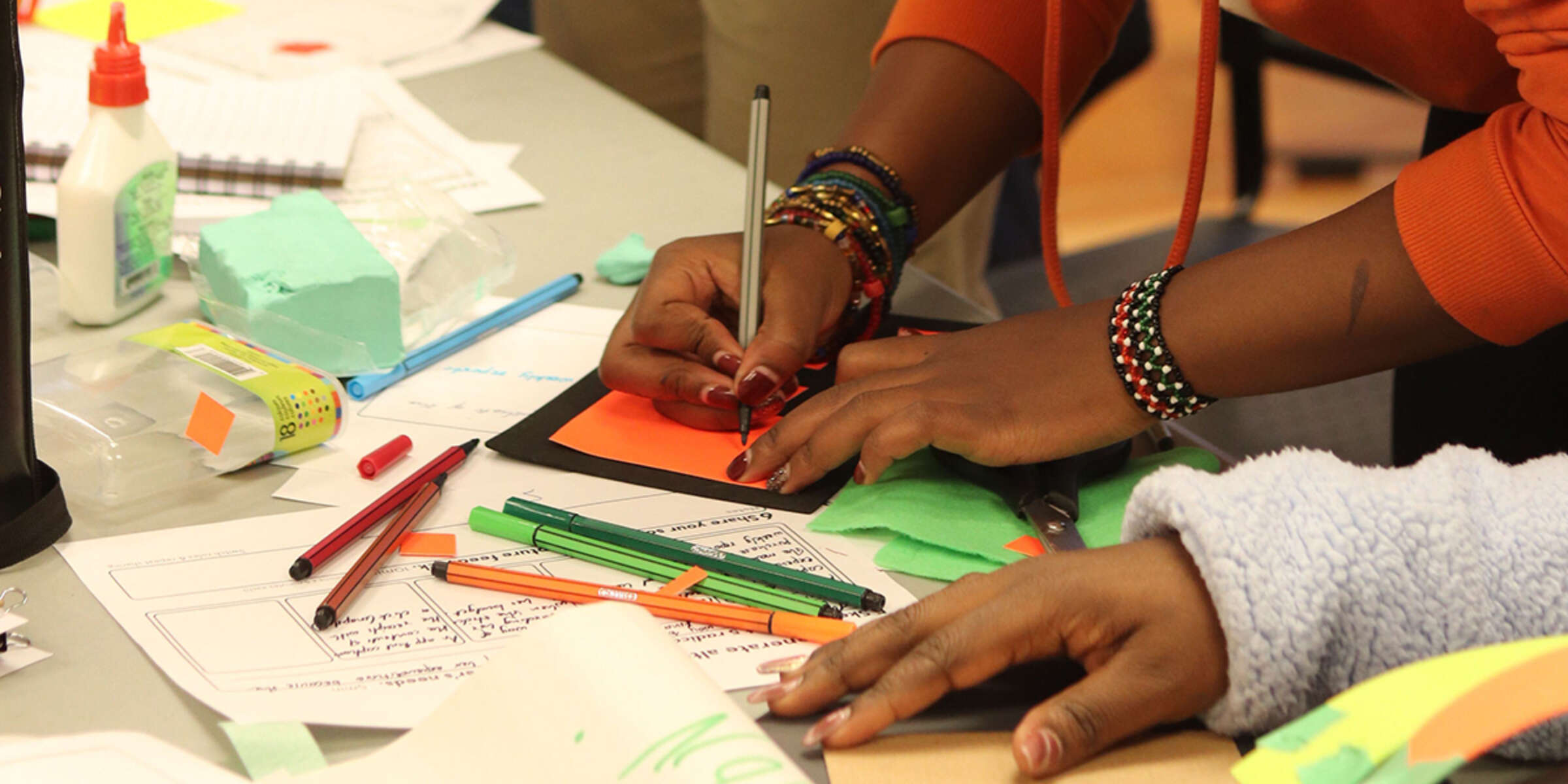 Someone is using a silver pen to draw on an orange piece of paper, surrounded by various art supplies like colored markers, glue, paper, and scissors on a table. The person is wearing multiple colorful bracelets and a red sweater. Another person's hand at the bottom of the image is holding paperwork.