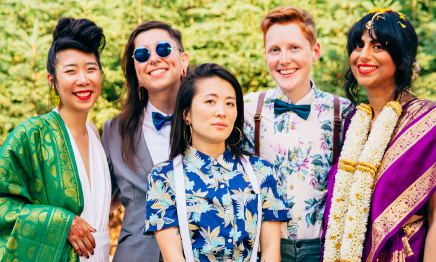 A group of five people standing outdoors in front of greenery. They are smiling and dressed in a variety of colorful outfits, including floral shirts, a gray suit, and traditional attire with garlands. One person is wearing sunglasses.