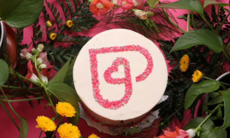 A round, white cake sits on a pink surface, surrounded by a variety of flowers and greenery. The cake is decorated with a red sugar outline of an abstract heart shape on top. Yellow and orange blooms are prominent among the surrounding foliage.