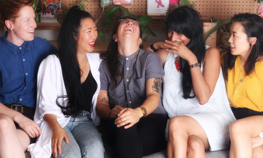 A group of five women sitting on a couch, smiling and laughing together. They are dressed in casual clothing and appear to be sharing a joyful moment. The background features shelves with books and plants.