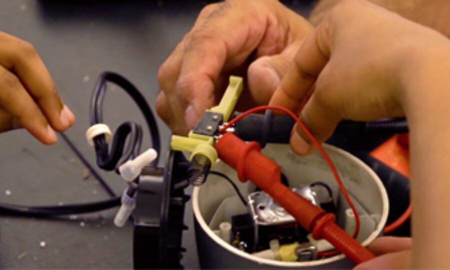 A close-up of a hands-on electronics project. Several hands are working together to assemble components including wires, a circuit board, and a switch, inside a small container. Tools and wires can be seen on the table around the project.