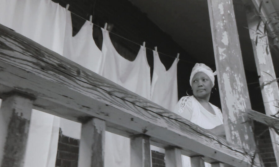 A black-and-white photo of a woman standing on a wooden porch. She looks pensively to the side, wearing a headscarf and a white top. Behind her, white sheets hang on a clothesline. The porch railings show signs of wear and age.