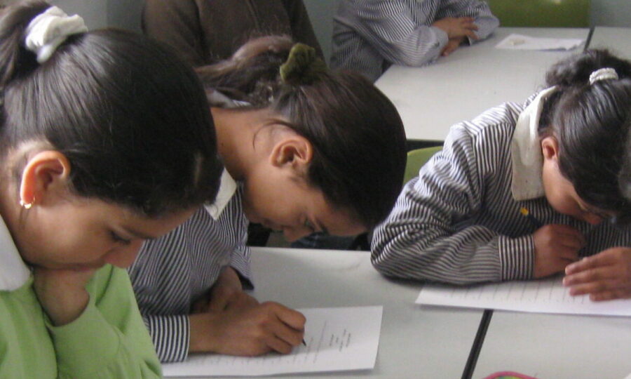 Three children with dark hair are sitting at a white table, leaning over their papers and writing with concentration. They are wearing striped uniforms. The background shows another student who is not in focus, seated at the same table.