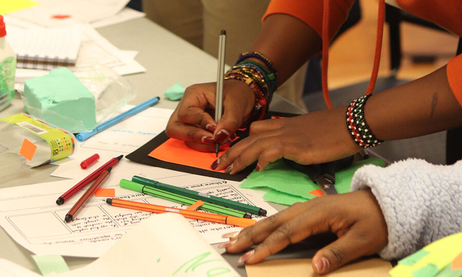 Someone is using a silver pen to draw on an orange piece of paper, surrounded by various art supplies like colored markers, glue, paper, and scissors on a table. The person is wearing multiple colorful bracelets and a red sweater. Another person's hand at the bottom of the image is holding paperwork.