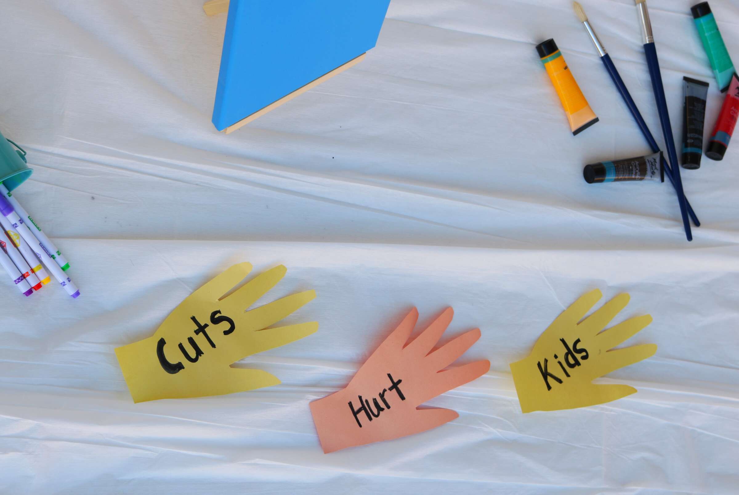 A table with art supplies, including paintbrushes, paint tubes, and markers, has three cut-out paper hands lying on it. The paper hands are yellow, orange, and pink, with the words "Cuts Hurt Kids" written on them.