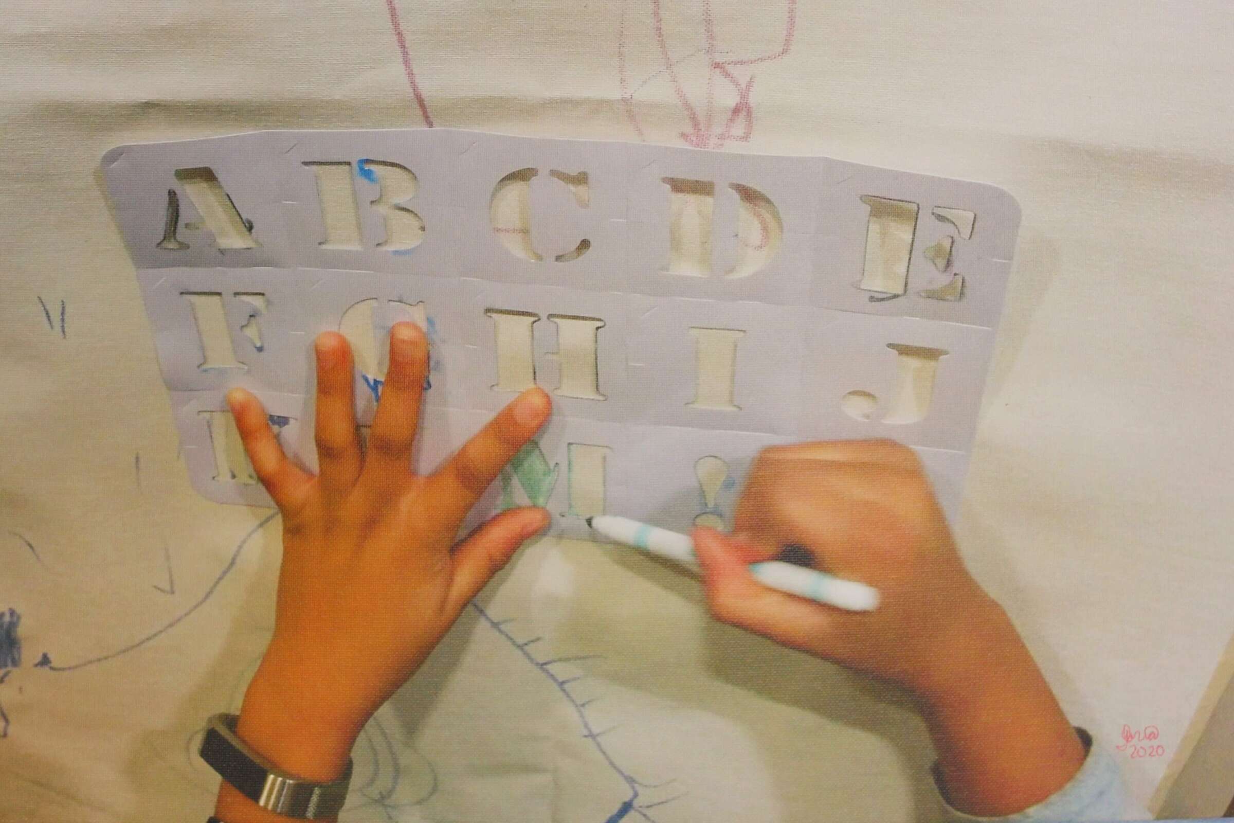 A child uses a stencil to trace uppercase letters of the alphabet on a large piece of paper. The child is holding a marker in one hand and stabilizing the stencil with the other. The letters "A-I" are visible, and some drawings are faintly seen on the paper.