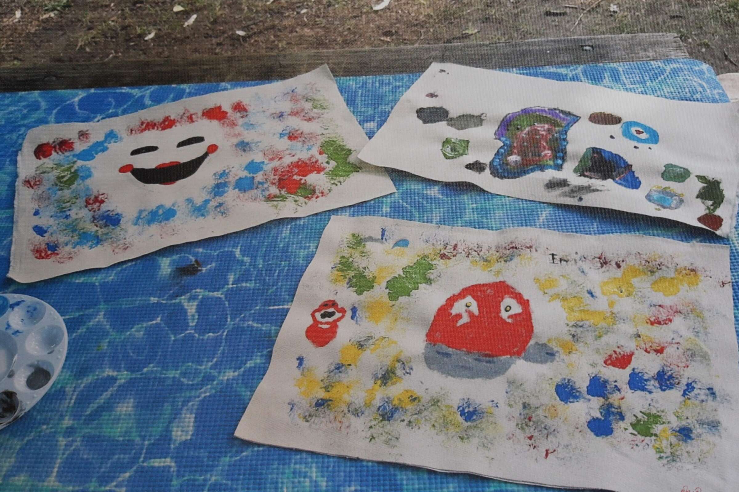 Three children's paintings on paper are laid out on a blue patterned tablecloth. The paintings feature colorful abstract designs, with one prominently showing a smiling face, another with various shapes and colors, and the third featuring patterns of red, blue, and yellow.