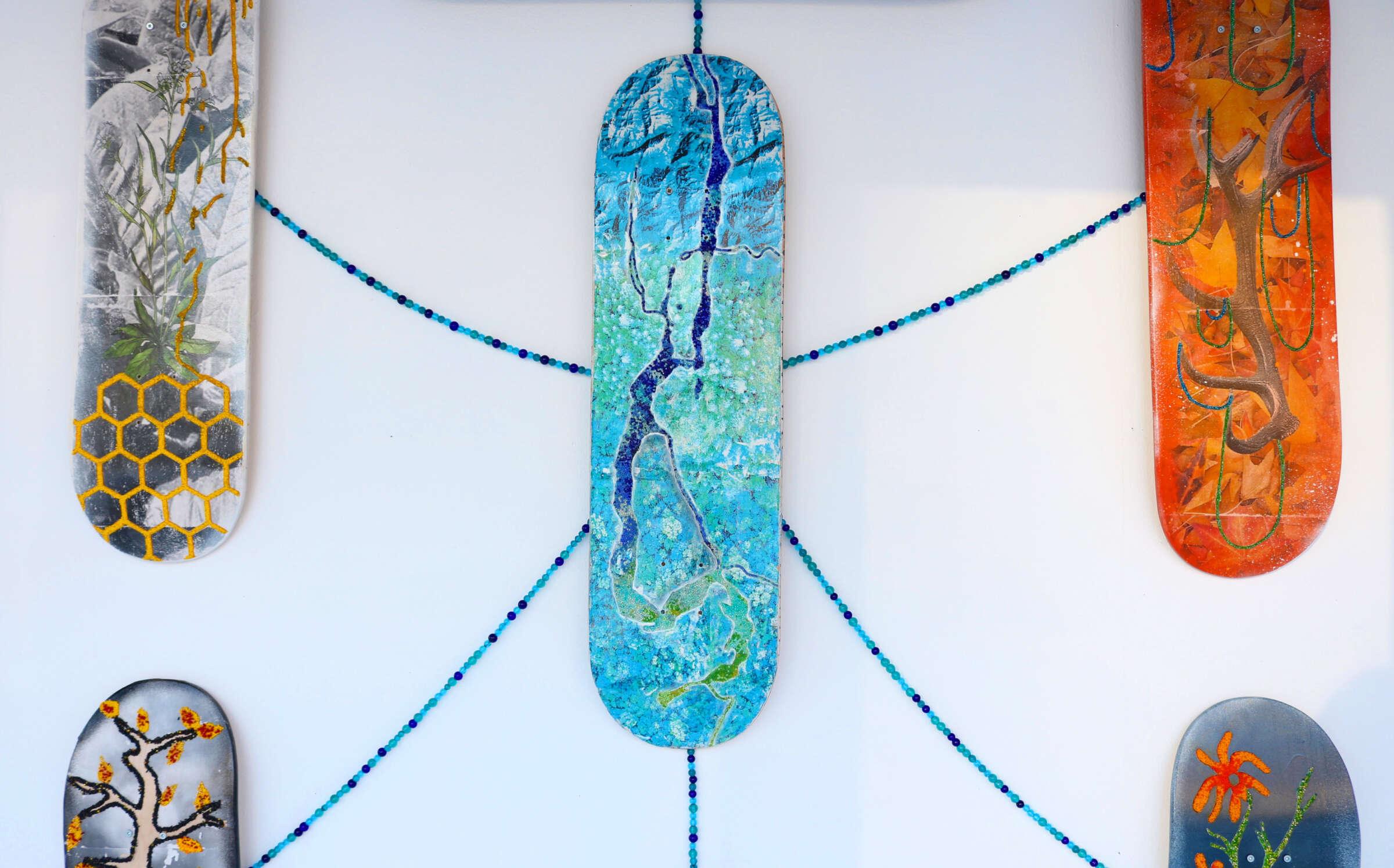 A collection of five vertical skateboards displayed on a wall. Each skateboard is uniquely decorated: one with a tree and octagonal patterns, one with a plant design, one with a blue river scene, one with an abstract tree, and one with red flowers. Blue beads connect the boards.