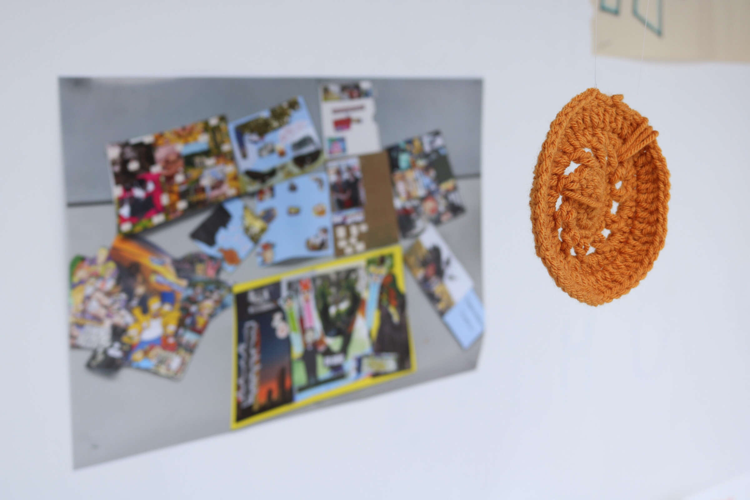 A crocheted, circular yellow ornament hangs in the foreground. Behind it on a wall, a collage of colorful images and papers is partially visible, displayed on a grey board. The focus is on the crocheted item, with the collage slightly blurred in the background.