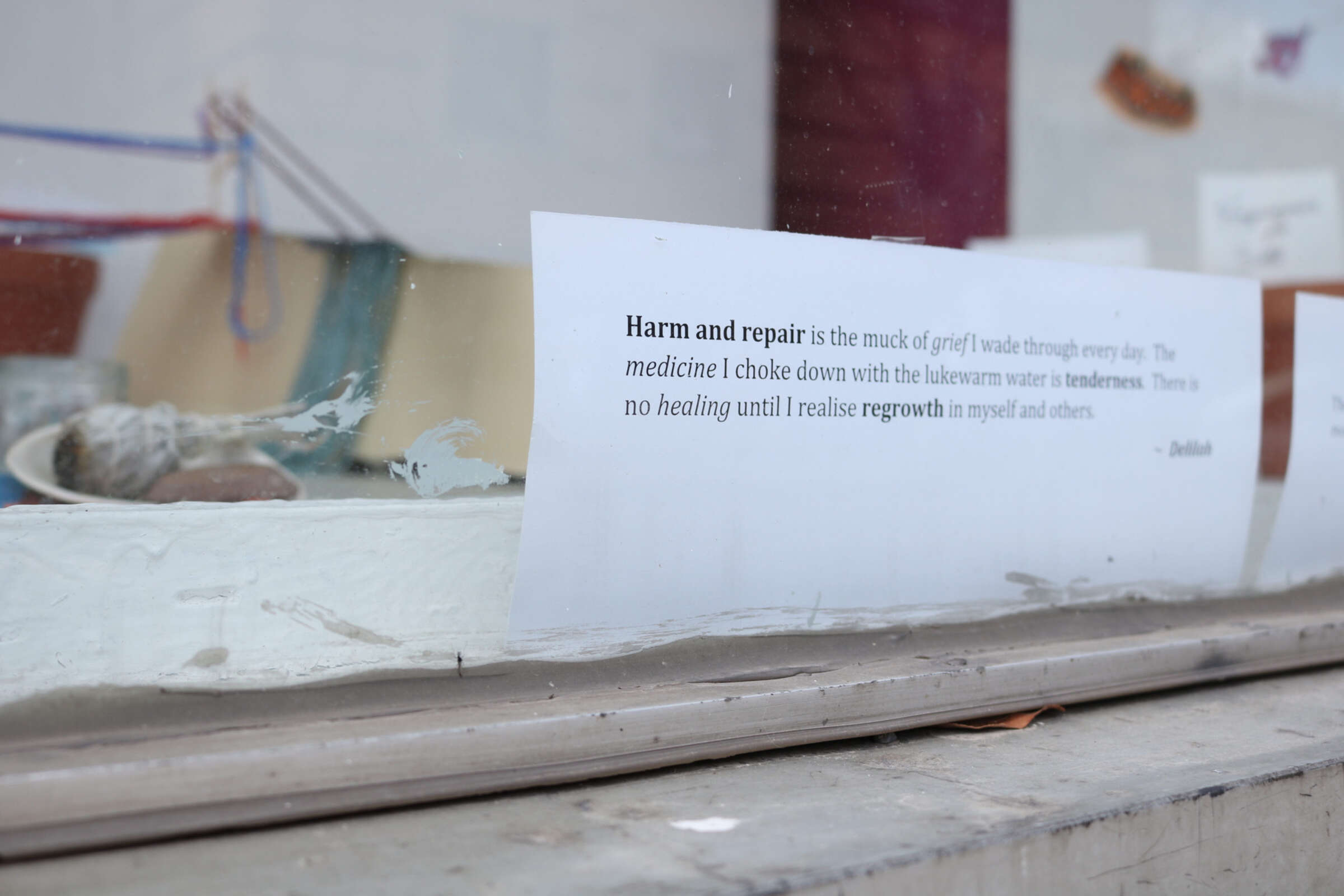 A close-up of a white paper with text taped to a glass window. The text reads, "Harm and repair is the muck of grief I wade through every day. The medicine I choke down with the lukewarm water is tenderness. There's no healing until I realise regrowth in myself and others. - Delilah." Blurred objects are visible in the background.