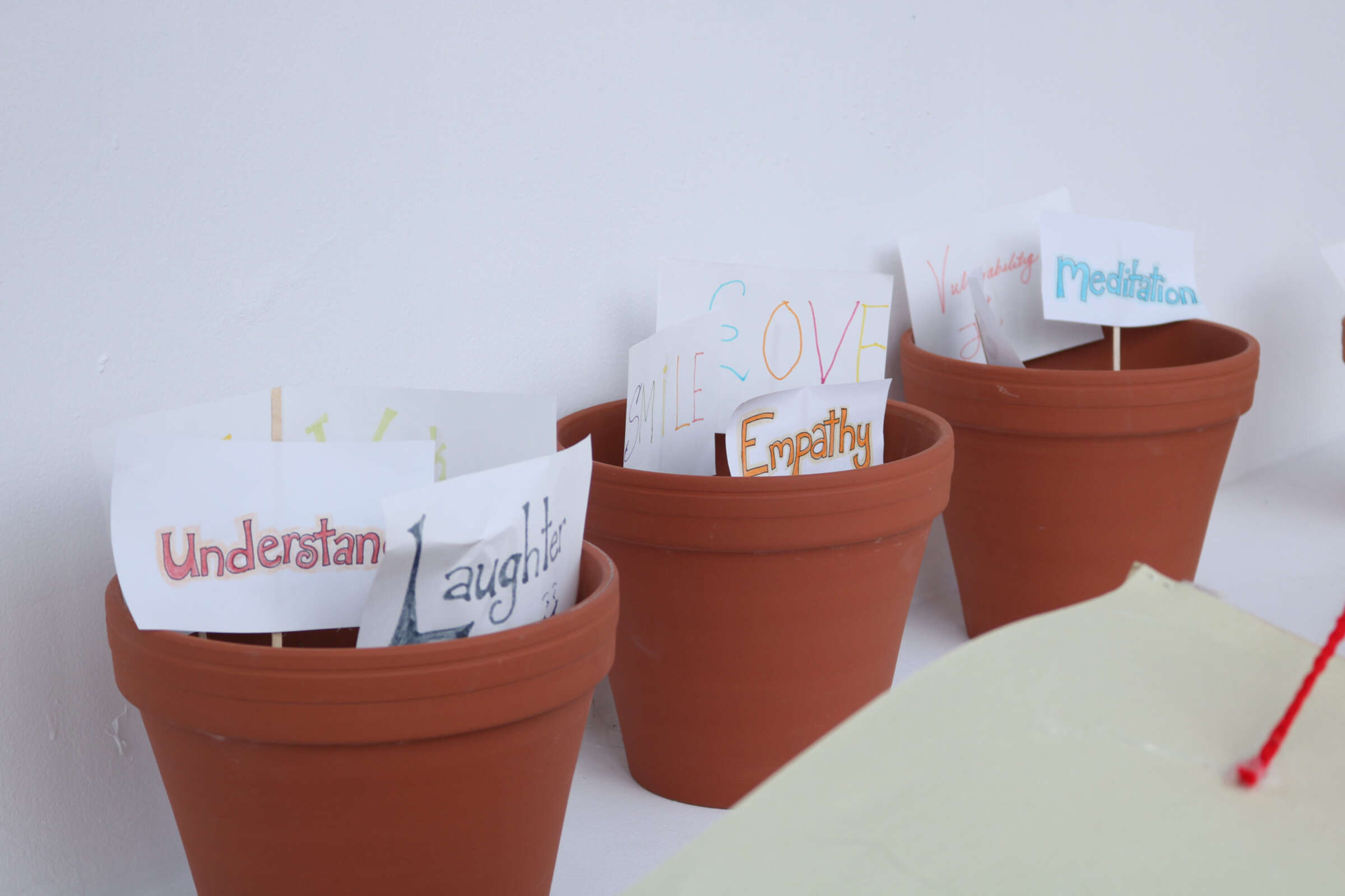 Four terracotta pots sit on a white surface, each containing handwritten signs with various positive messages. Some text includes "Laugh," "Understand," "Empathy," and "Meditation." The signs are colorful and creatively designed.