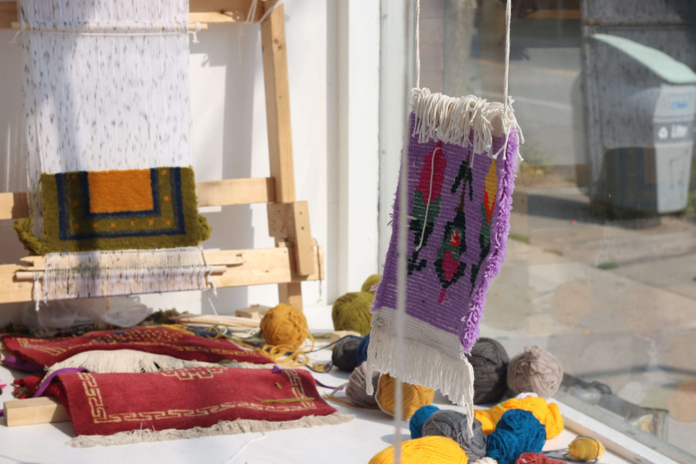 A workshop table displays handmade woven textiles, yarn balls in various colors, and some tools. A tapestry with a purple border and intricate patterns is hanging in the foreground, while other colorful weavings and a loom are visible in the background.