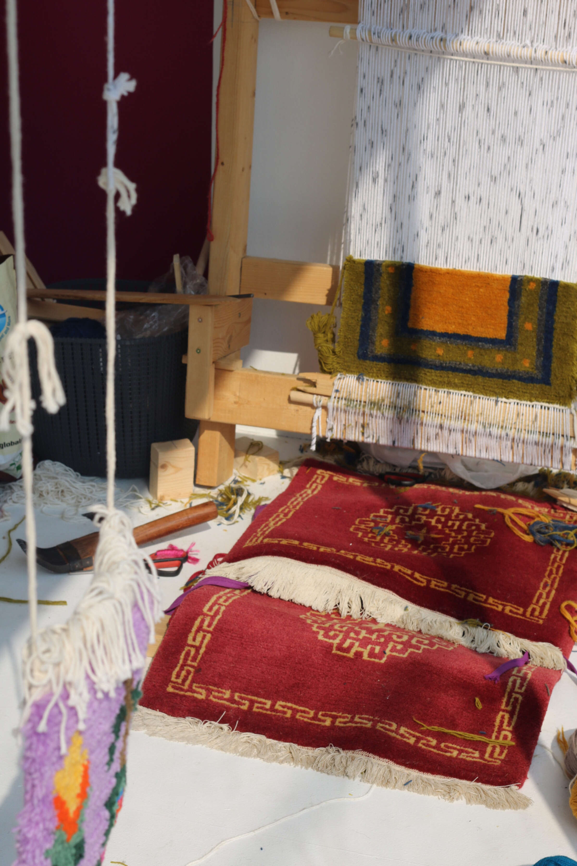 A weaving loom with partially completed textiles is surrounded by vibrant, handmade rugs and weaving materials. The textiles feature intricate patterns in various colors, including red, yellow, and purple. Some tools and yarns are scattered around the workspace.