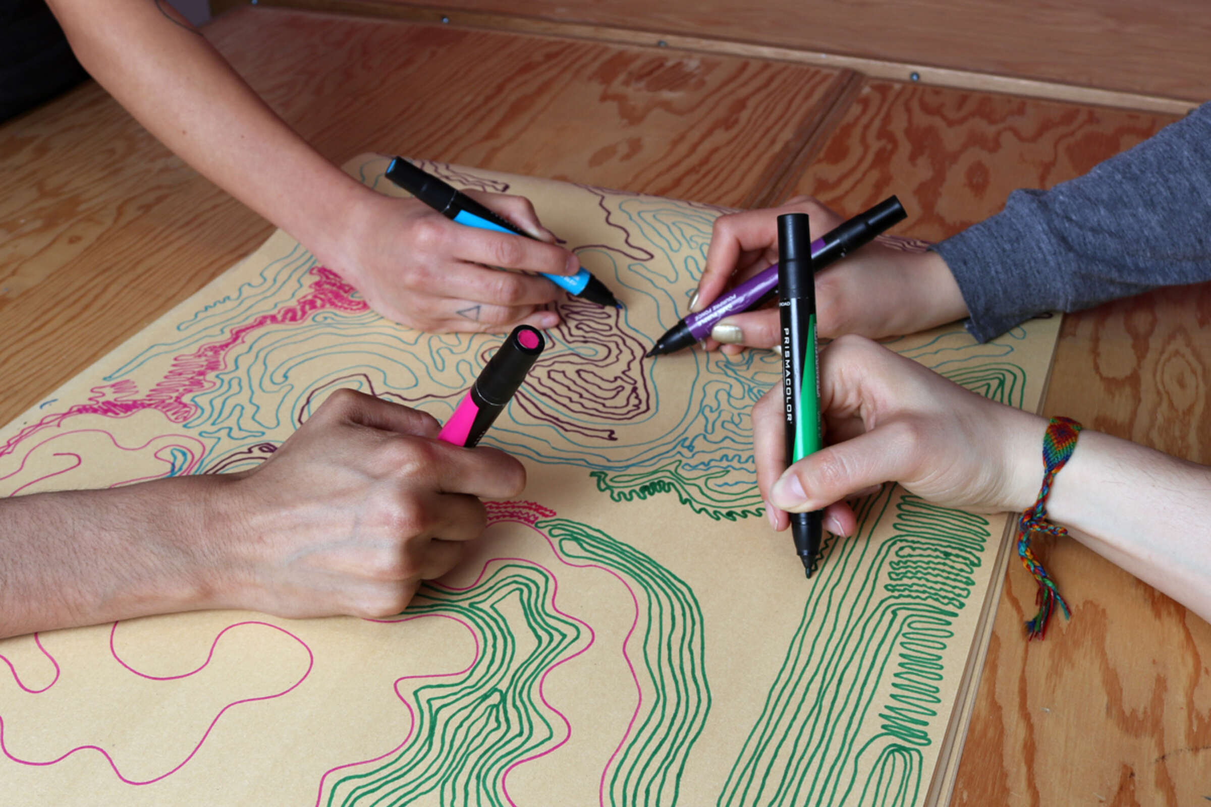 Four hands are collaboratively drawing on a large piece of paper with colorful markers on a wooden table. The paper displays various colorful lines and patterns. The hands are of diverse individuals, with varying skin tones and accessories.