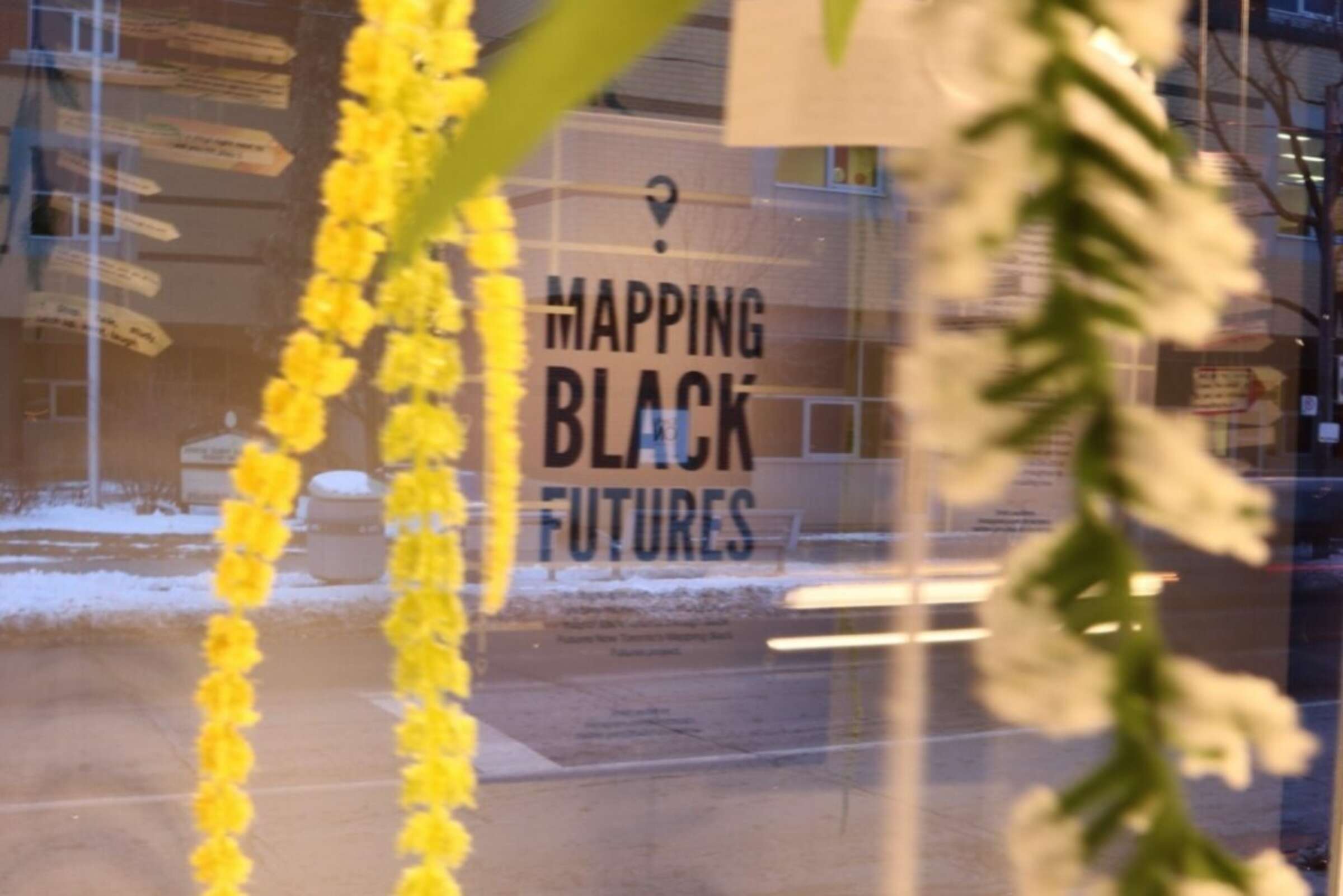 Yellow and white flower garlands are hanging in the foreground, partially obscuring a window with the text "Mapping Black Futures." A snowy street with buildings and trees can be seen through the window.