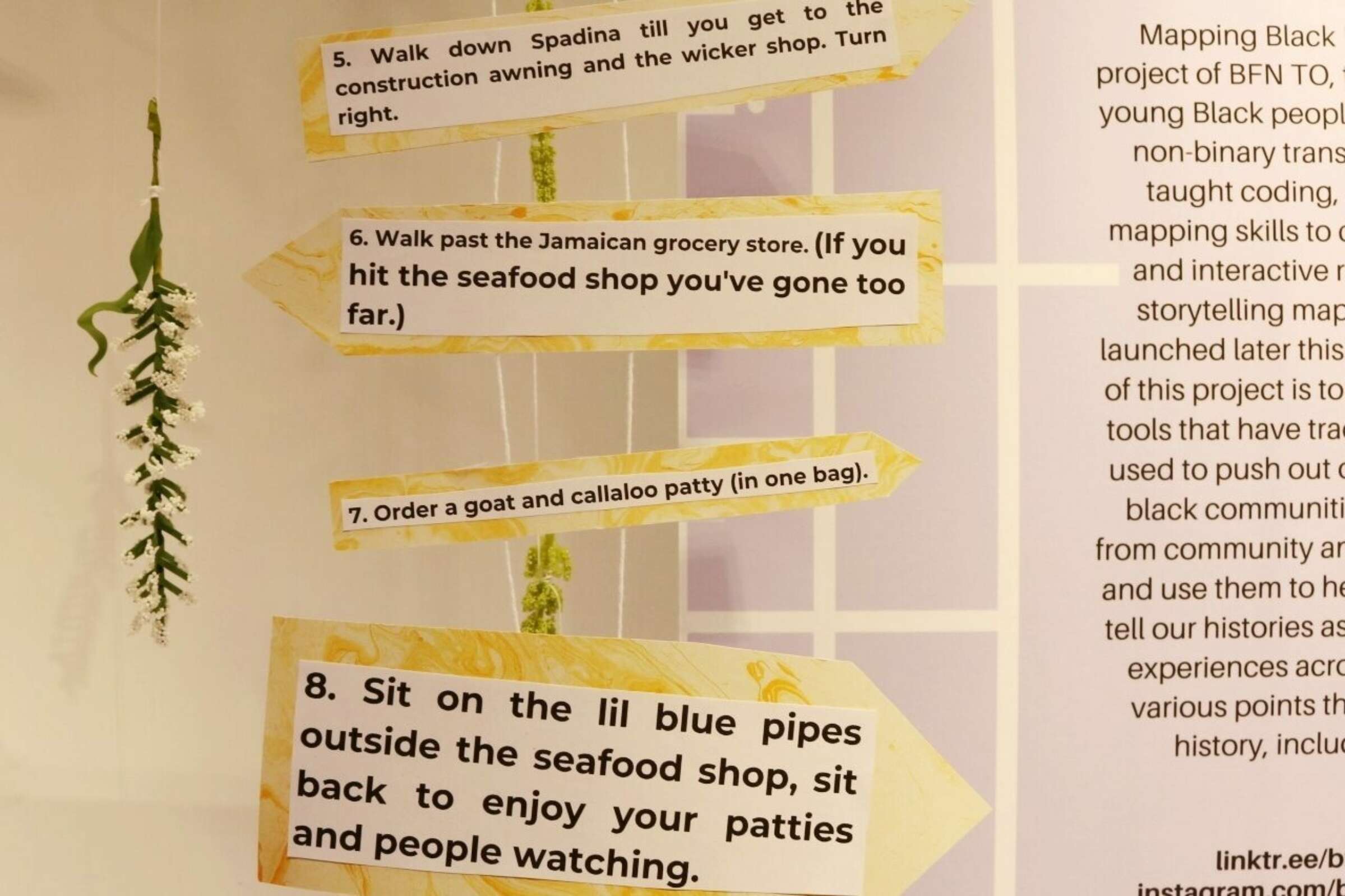 Three wooden signposts displaying instructions for a walking tour are hanging from a string. The numbered steps advise walking directions, noting landmarks like a Jamaican grocery store and a seafood shop, and suggest sitting on blue pipes to enjoy patties and people-watching.