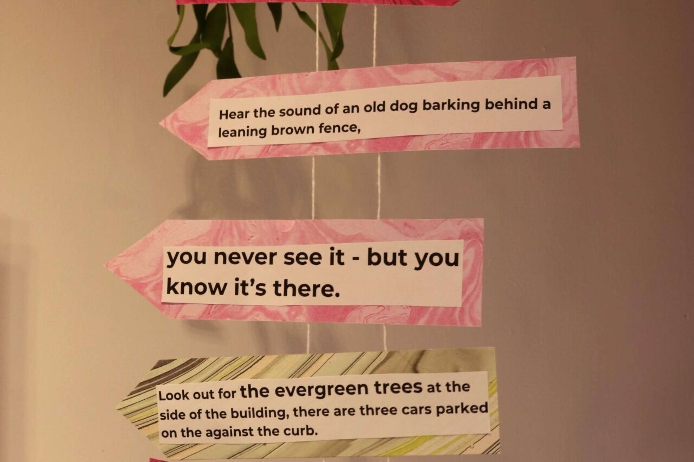 Three pink and yellow paper arrows with typed text are hanging against a beige wall. The texts on the arrows are: "Hear the sound of an old dog barking behind a leaning brown fence," "you never see it - but you know it’s there," and "Look out for the evergreen trees at the side of the building, there are three cars parked on the against the curb.