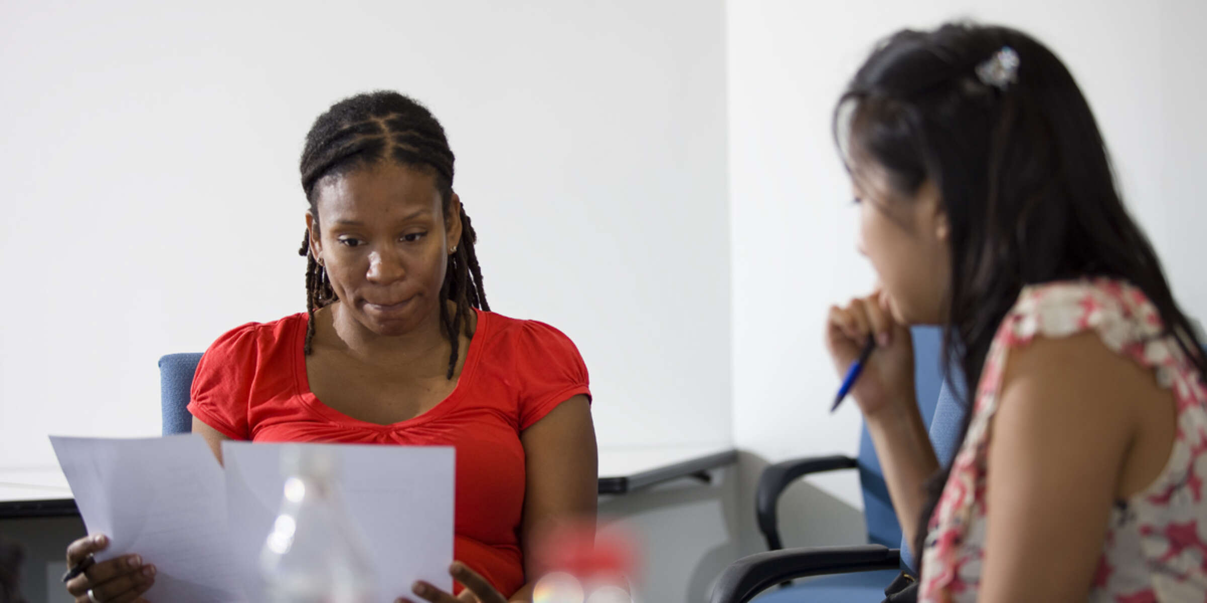 Two women seated at a table. The woman on the left, in a red top, is reading sheets of paper with a thoughtful expression. The woman on the right, in a patterned top, is holding a pen and appears to be speaking. Both are in a light-colored, modern office setting.