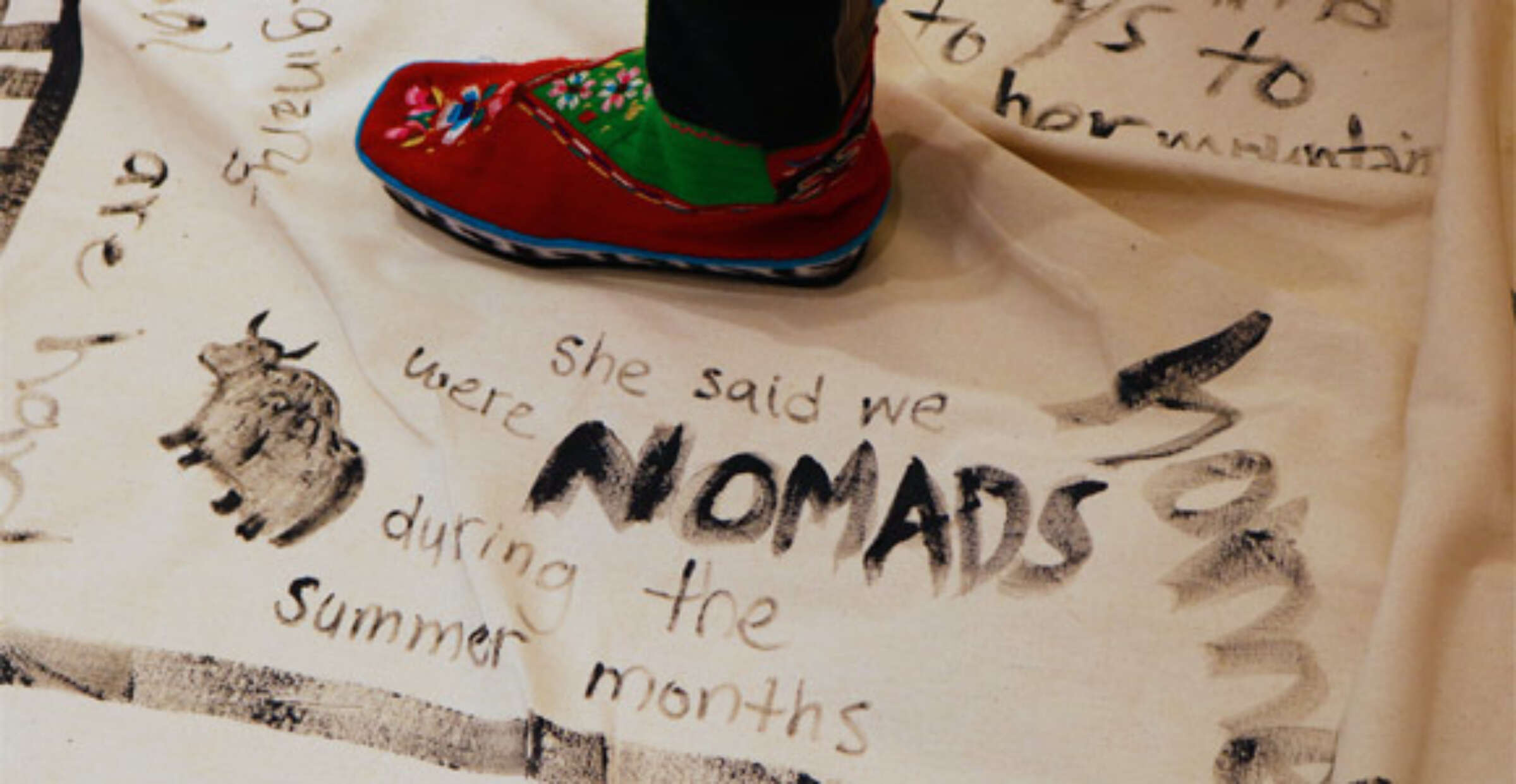 A person's foot wearing a red and green shoe stands on a beige cloth. The cloth has drawings and handwritten text, including the phrases "she said we were NOMADS during the summer months" and illustrations of a yak and partial, indistinct words.