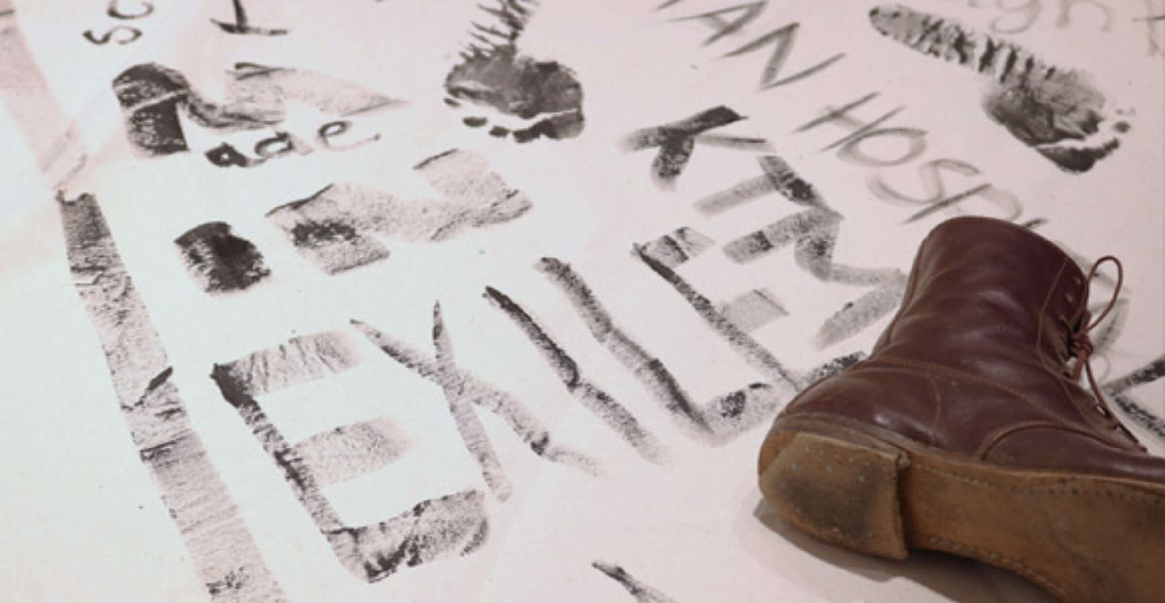 A close-up of writing and footprints in black paint on a white surface. The word "EXILES" is prominently visible among the markings. A brown boot is partially visible in the corner of the image, suggesting movement or recent activity.