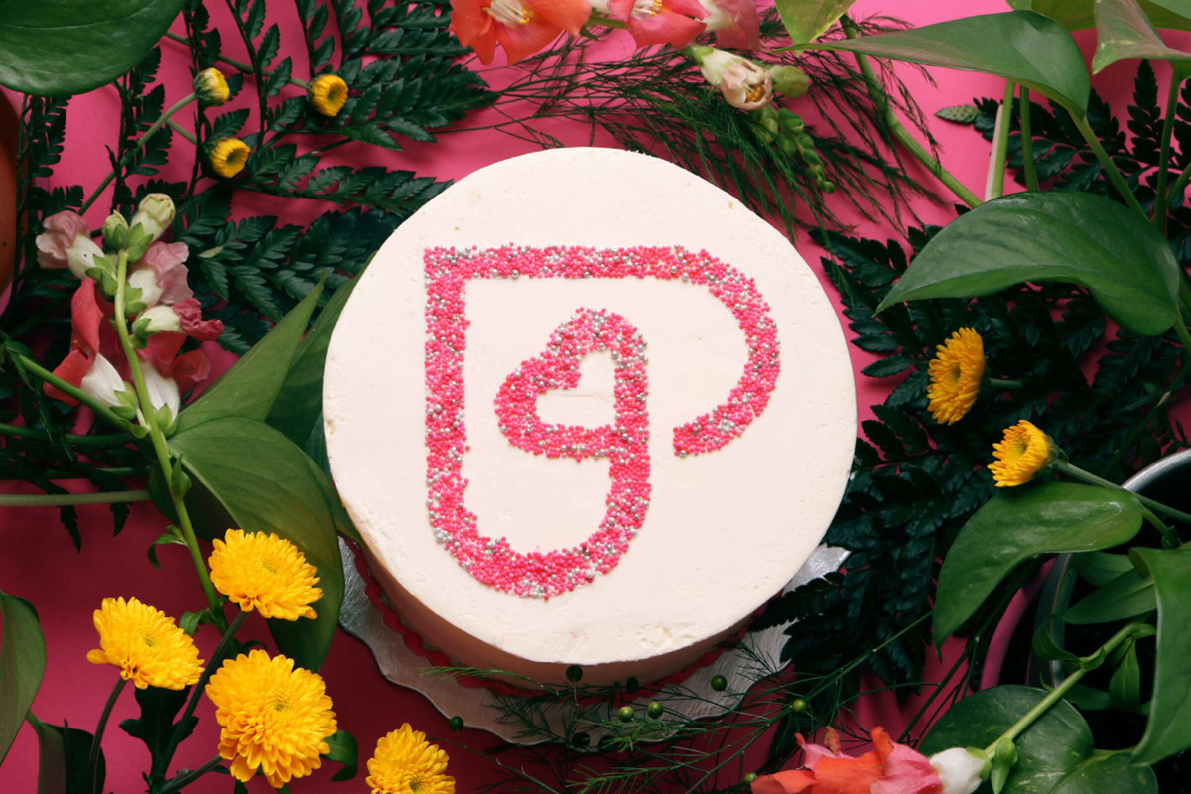 A round cake sits on a pink surface, surrounded by an arrangement of vibrant flowers and green foliage. The cake is frosted with white icing and decorated with a red heart design made from sprinkles in the center.