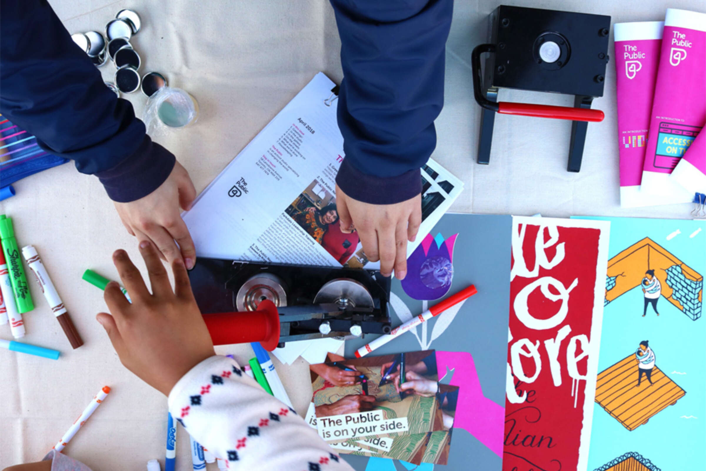Top-down view of a table filled with art supplies and papers. Several hands are actively engaged in cutting, arranging, and working on colorful projects. The table features markers, scissors, tape, pamphlets, and craft materials.