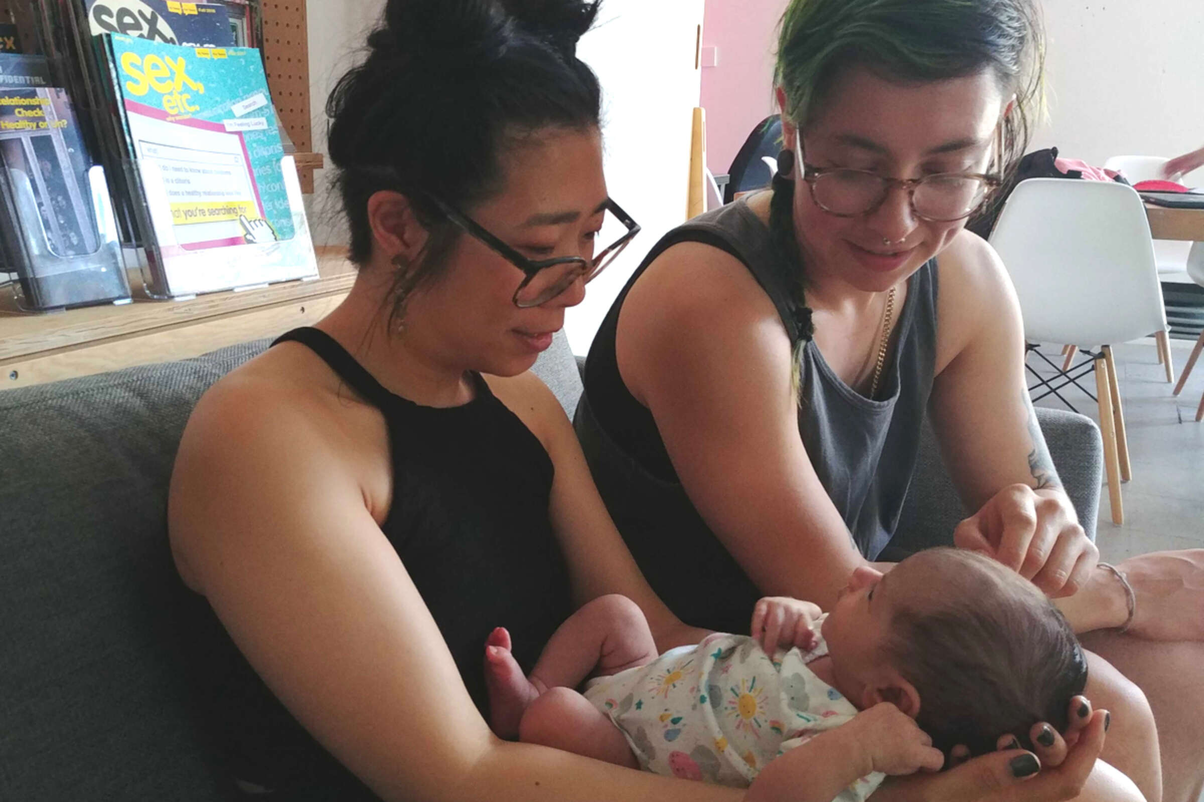 Two adults with glasses are sitting on a couch, lovingly looking at a baby they're holding. The person on the left in a black tank top cradles the baby, while the person on the right in a gray top gently touches the baby's head. Shelves with magazines are in the background.