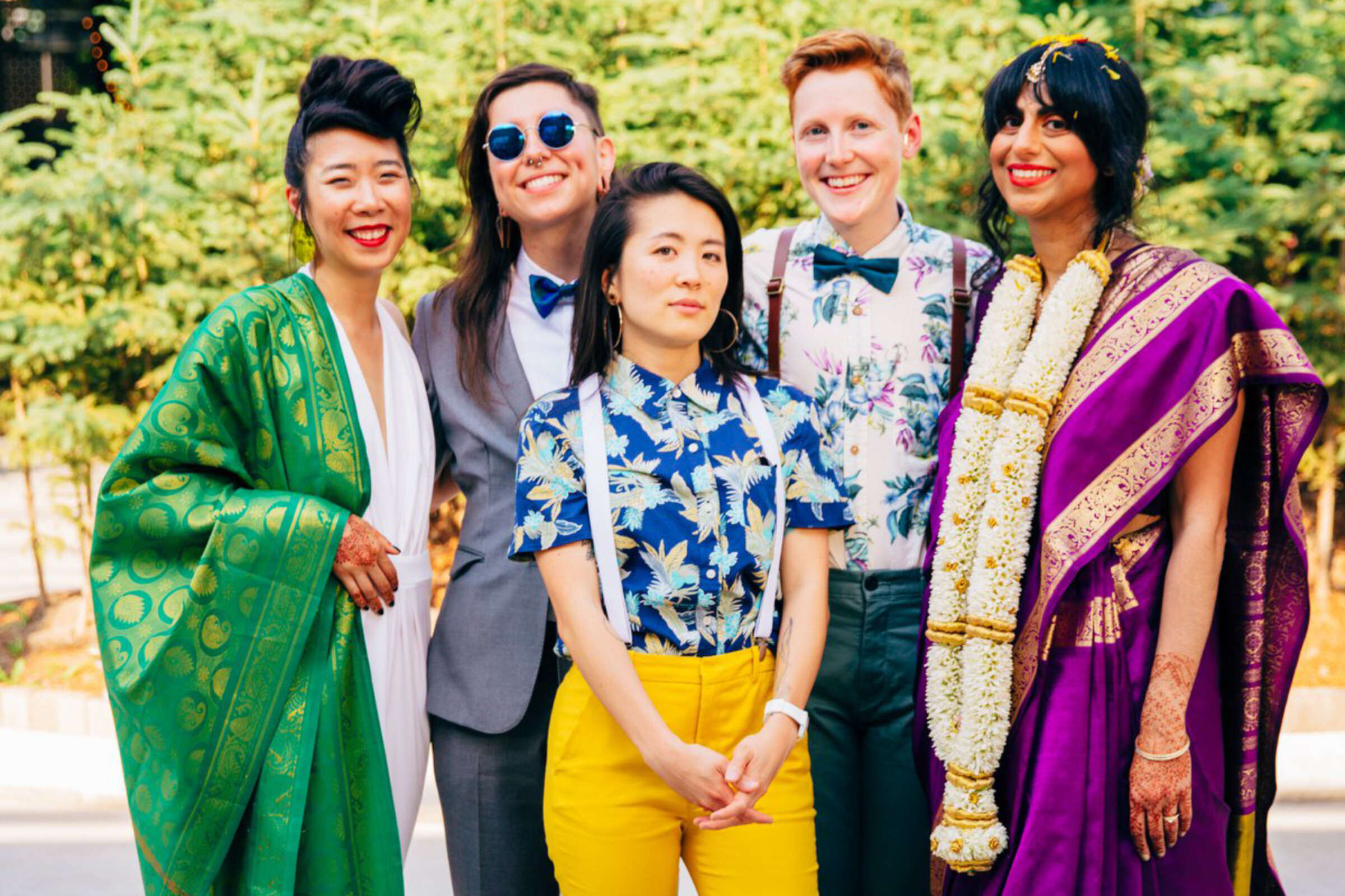 Five people pose together outdoors, smiling at the camera. They are dressed in a variety of colorful, eclectic outfits, including a green robe, a floral shirt with yellow pants, a purple sari with gold trim, and a suit with a bow tie. Trees are visible in the background.