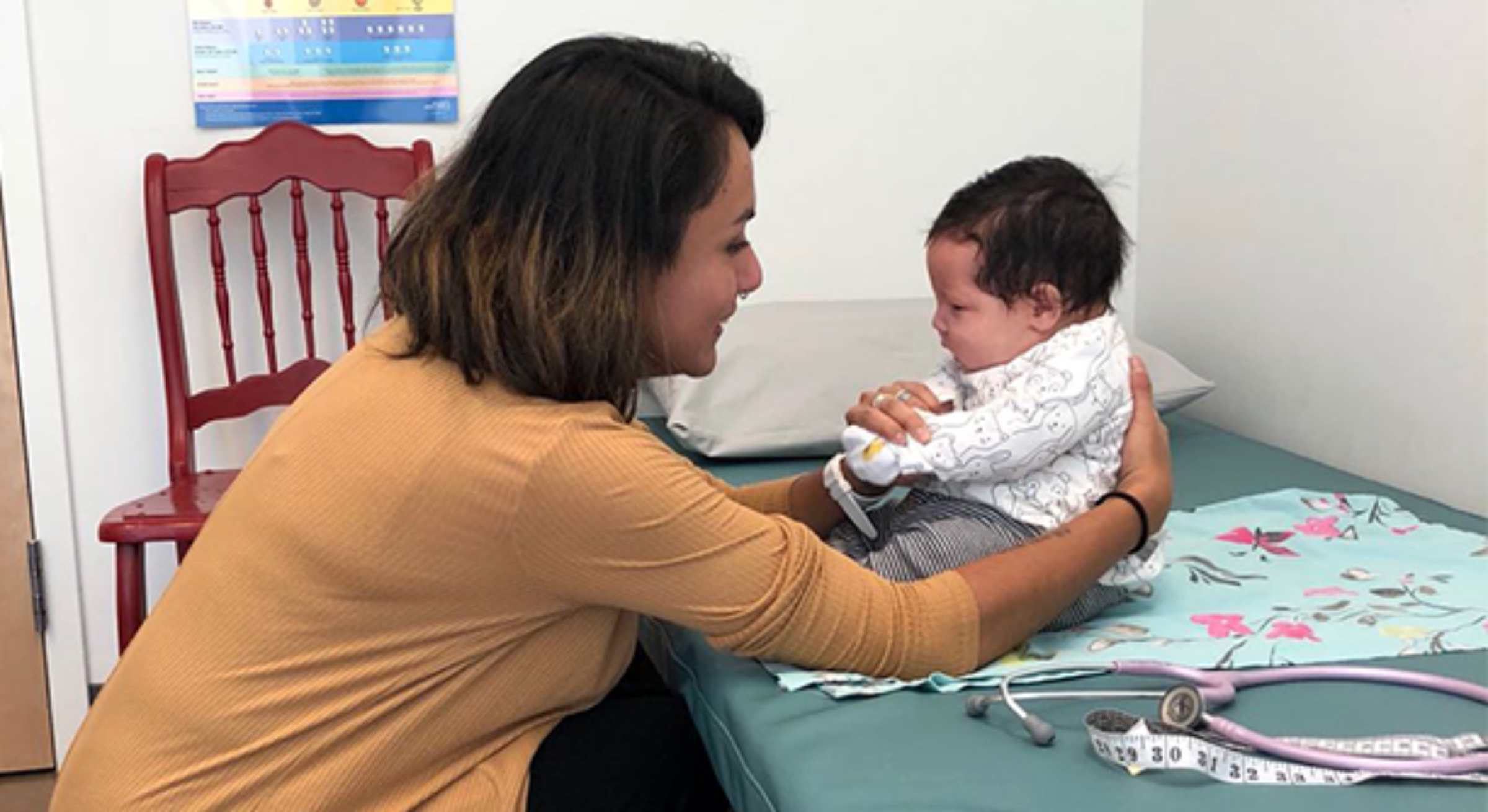 A woman in a mustard yellow shirt is sitting on the edge of a medical examination table, holding a baby dressed in white and patterned clothing. The baby sits on the table with a measuring tape and a stethoscope nearby. A red chair and wall chart are visible in the background.