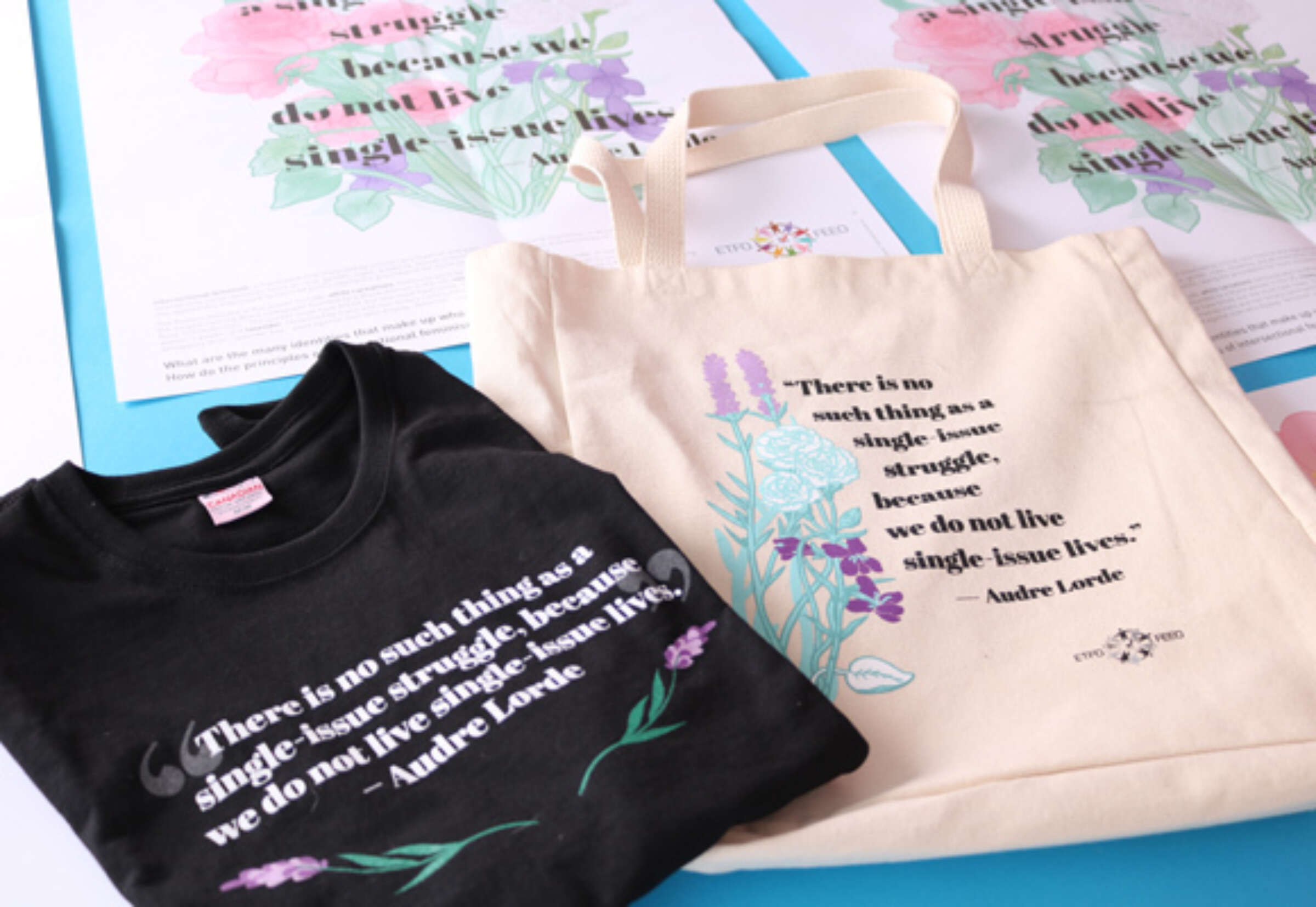 Displayed on a blue surface are a black T-shirt and a beige tote bag, both featuring a quote by Audre Lorde: "There is no such thing as a single-issue struggle because we do not live single-issue lives," and decorated with lavender and floral illustrations.