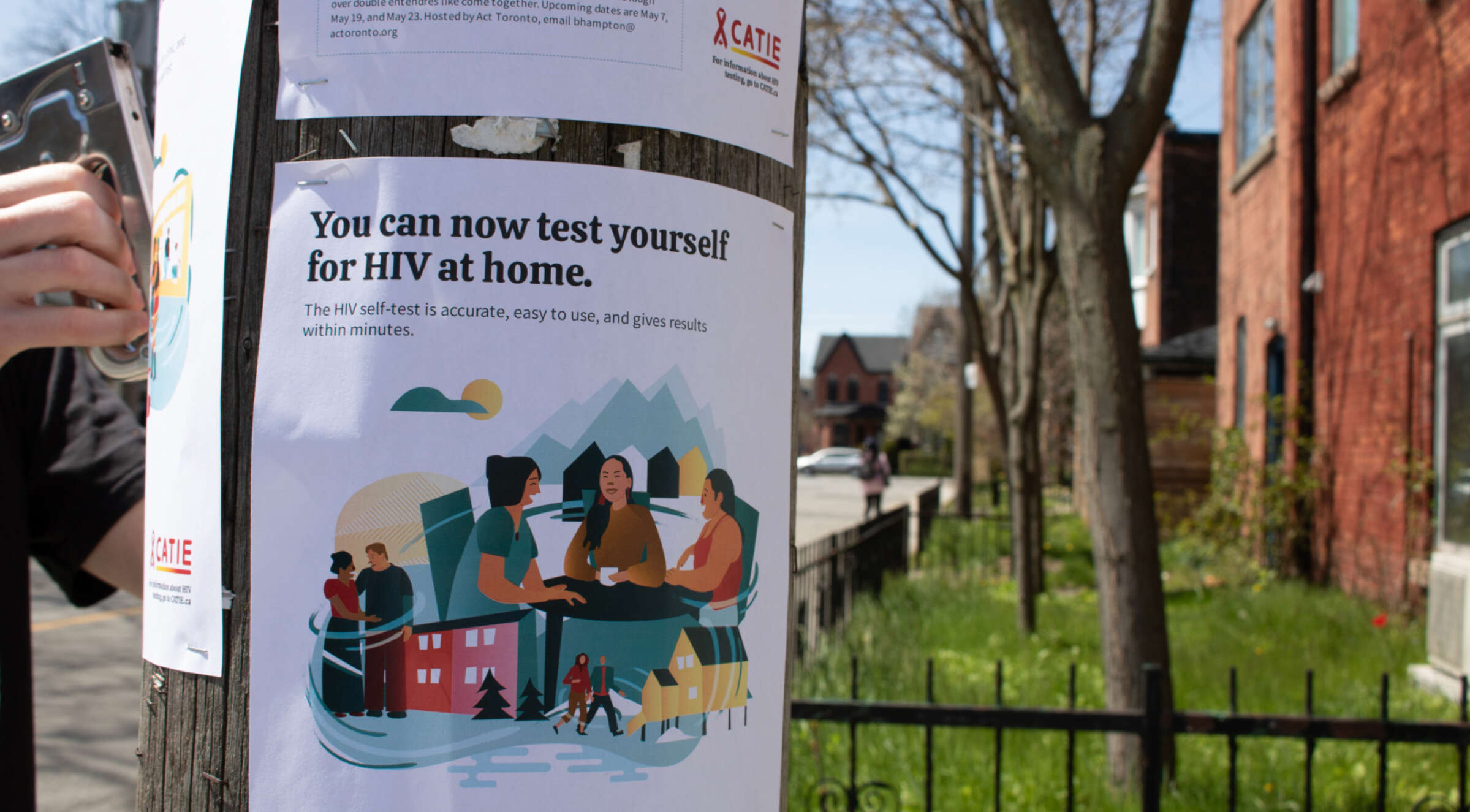 A person is putting up a poster on a wooden pole. The poster reads, "You can now test yourself for HIV at home. The self-test is accurate, easy to use, and gives results within minutes." It includes an illustration of people gathered together with an urban backdrop.