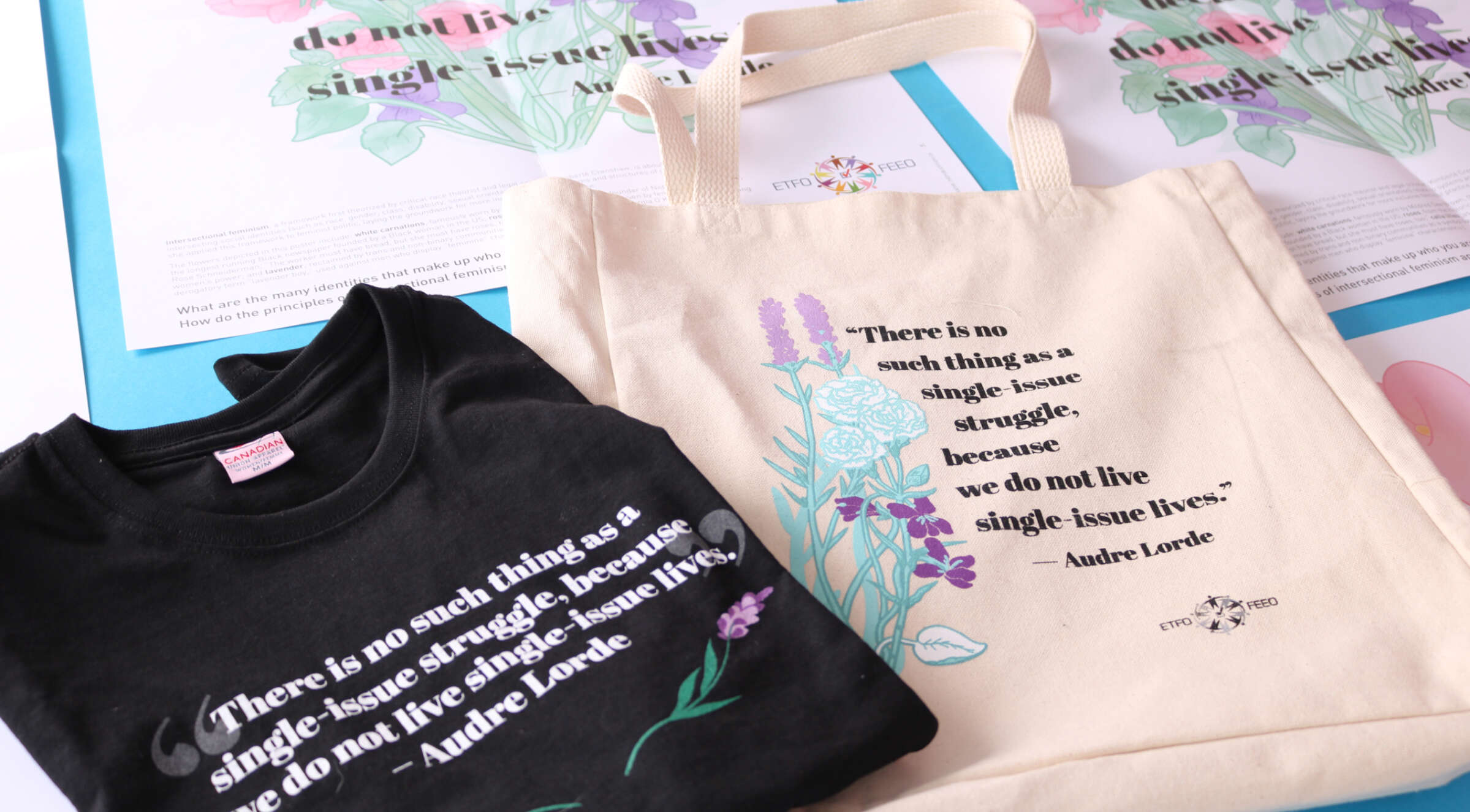 A black t-shirt and a cream-colored tote bag are displayed on a table. Both feature a quote by Audre Lorde, "There is no such thing as a single-issue struggle because we do not live single-issue lives," along with colorful floral illustrations.