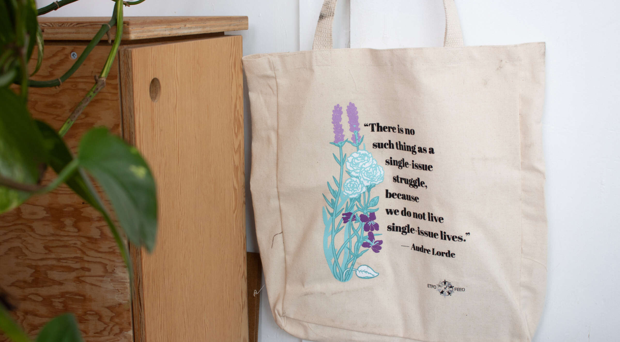 A cream-colored tote bag hangs on a wooden piece of furniture. The bag features an illustration of blue and purple flowers and the quote "There is no such thing as a single-issue struggle, because we do not live single-issue lives." - Audre Lorde. A green plant is partially visible on the left.