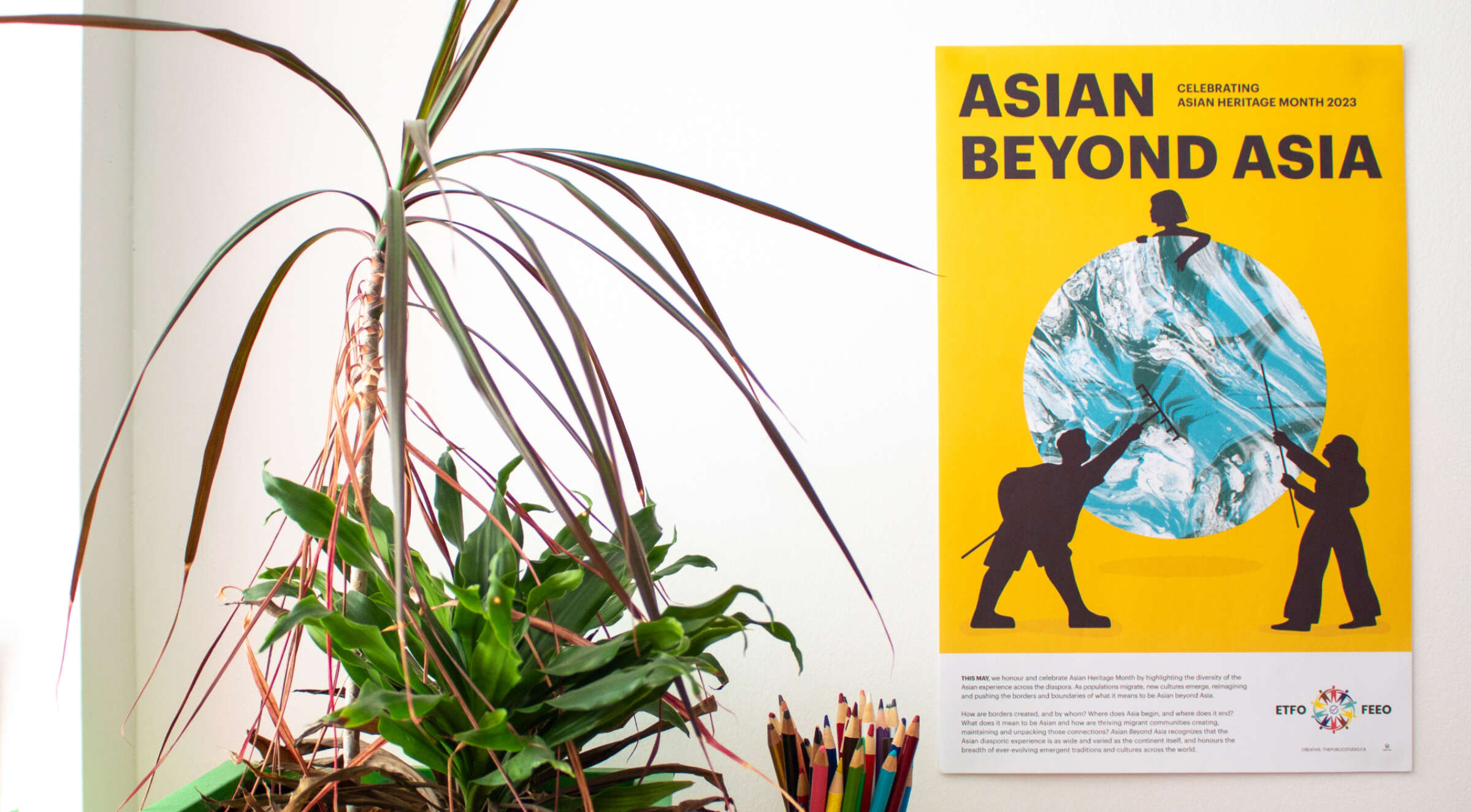 A vibrant yellow poster on a white wall reads "ASIAN BEYOND ASIA" and "Celebrating Asian Heritage Month 2023" with a globe and silhouettes of people. The poster is next to a tall potted plant, and a bunch of colored pencils is visible below the poster.