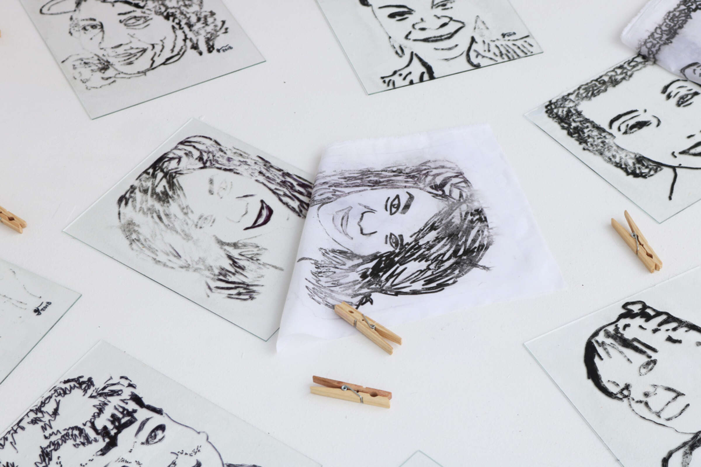 A collection of black and white sketches of faces placed on a white surface. Some sketches are on transparent sheets, and one sketch on a white cloth is being revealed. Wooden clothespins are scattered throughout the scene.