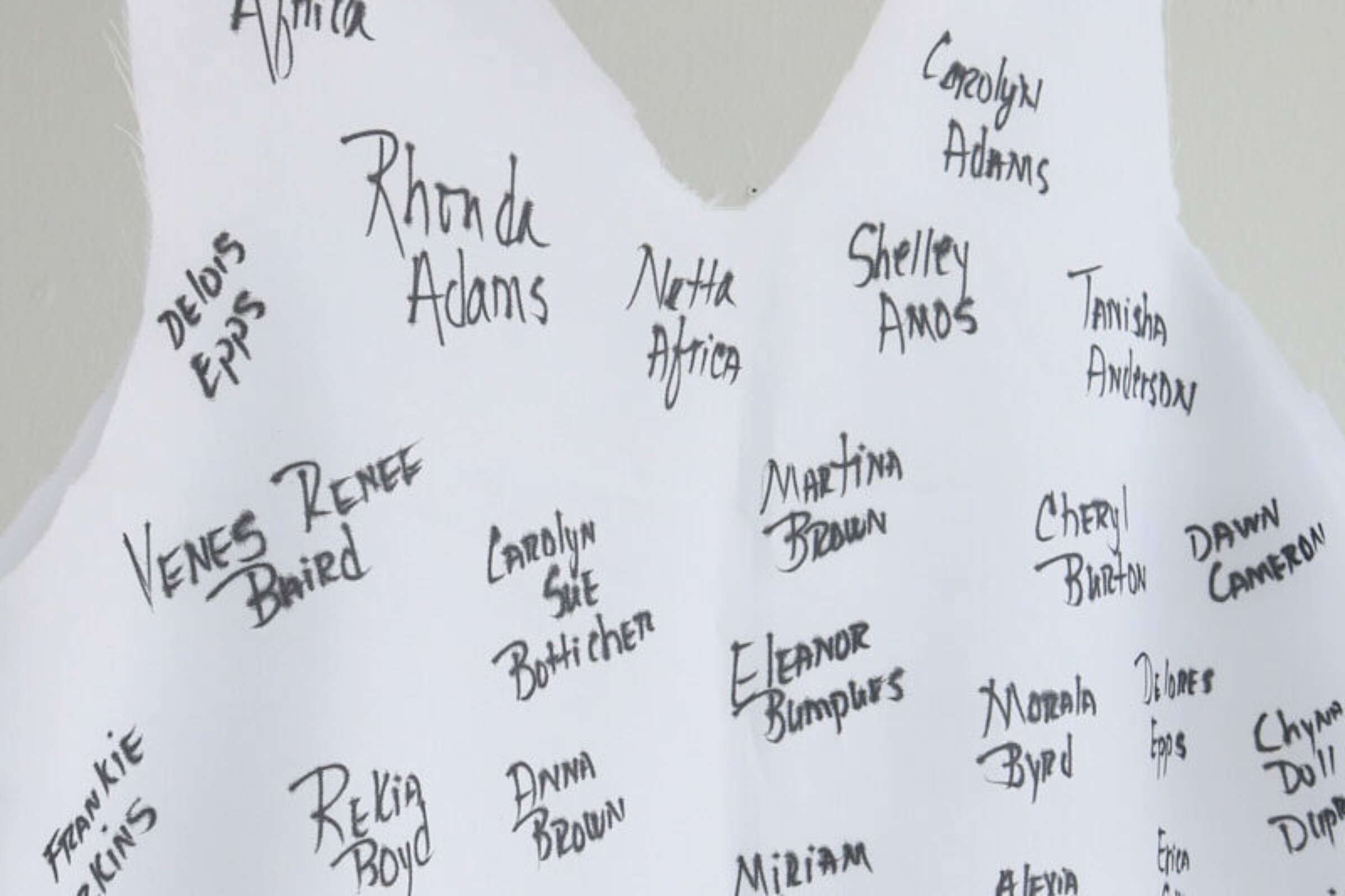 A close-up photo of a white paper dress with numerous handwritten names in black ink. The names are written in a random arrangement, creating a pattern across the surface of the dress. The names are written in various sizes and styles of handwriting.