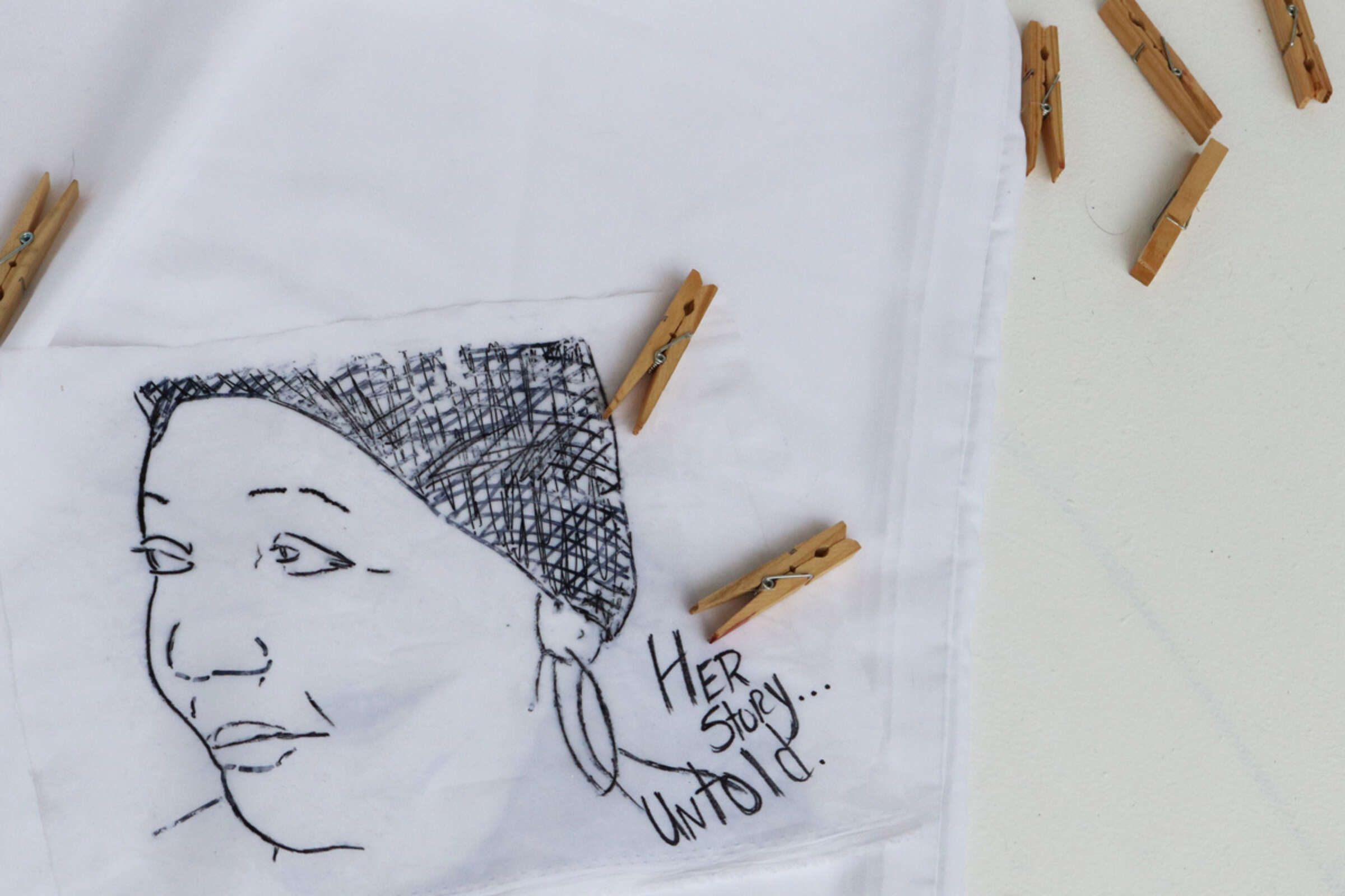 A piece of white fabric featuring a black ink drawn portrait of a woman wearing a headscarf and large hoop earrings. The fabric is pinned by wooden clothespins. Below the portrait are the handwritten words: "HER story UNTOLD...