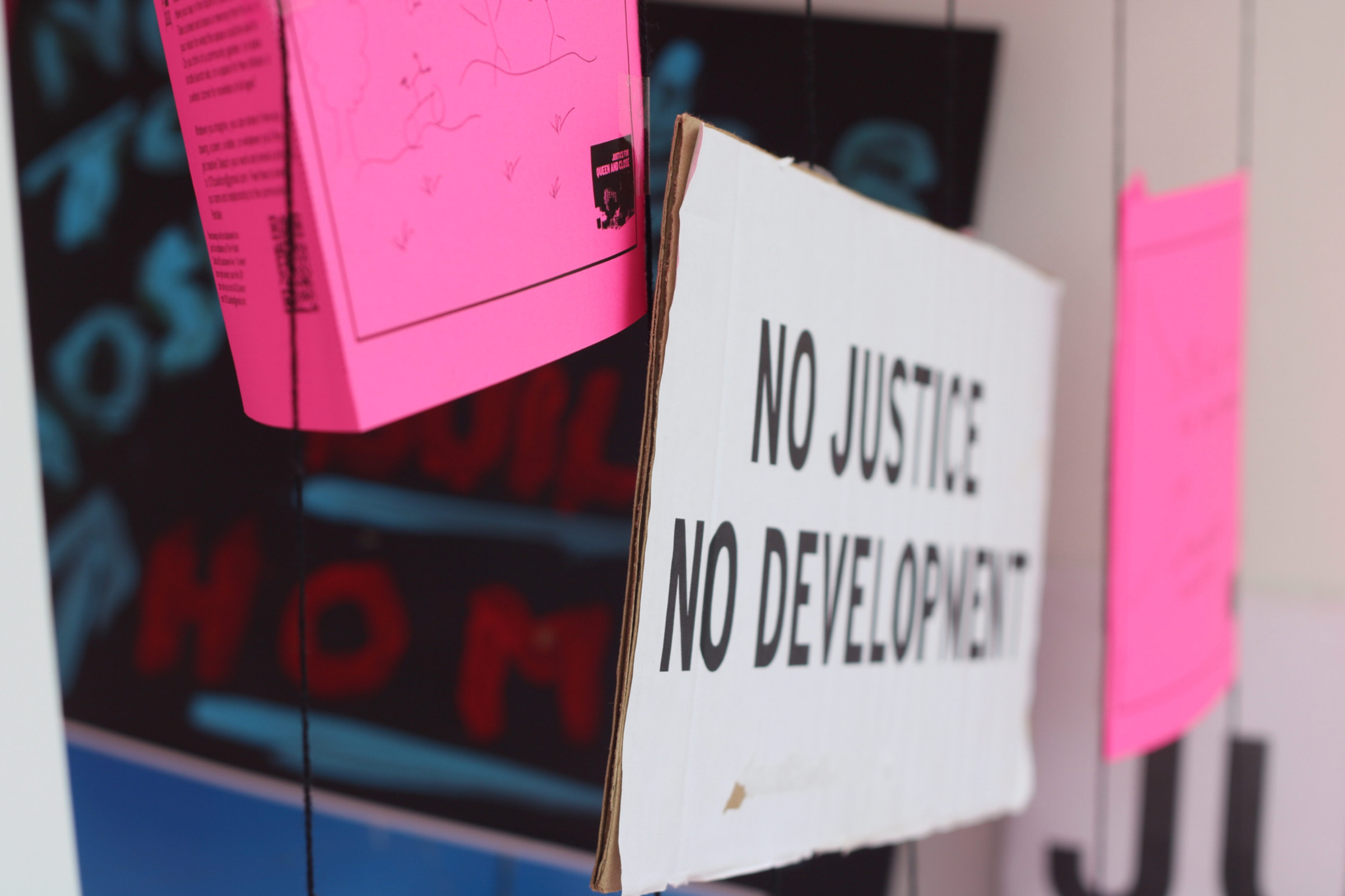 Close-up of protest signs hanging from strings. The central white sign reads, "NO JUSTICE, NO DEVELOPMENT." Other signs are visible in the background, including one pink sign with partially legible text. The image conveys a strong message about justice and development.