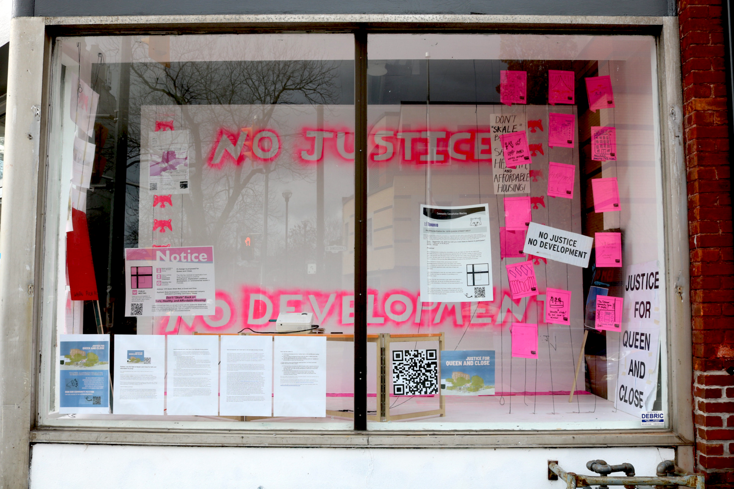 A storefront window displays multiple posters and notices, predominantly in bright pink. Neon text inside reads "No Justice, No Development." Additional signs include "Don't Make My Home A Monopolized Playground" and "Justice For Queen and Close.