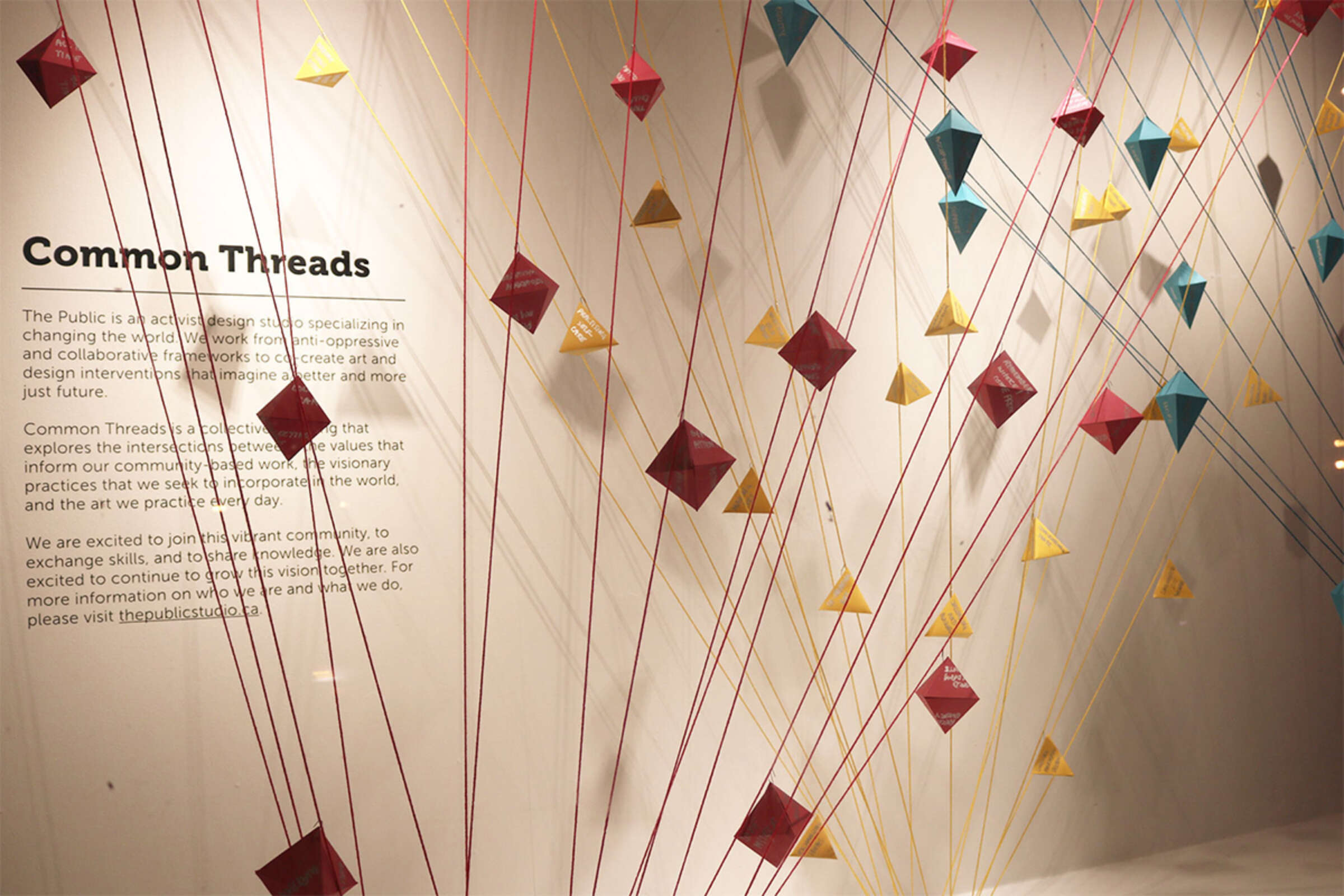 An art installation featuring colorful diamond-shaped pieces of fabric suspended by strings against a white background. The exhibit is titled "Common Threads," and a description of the artwork is written on the wall.