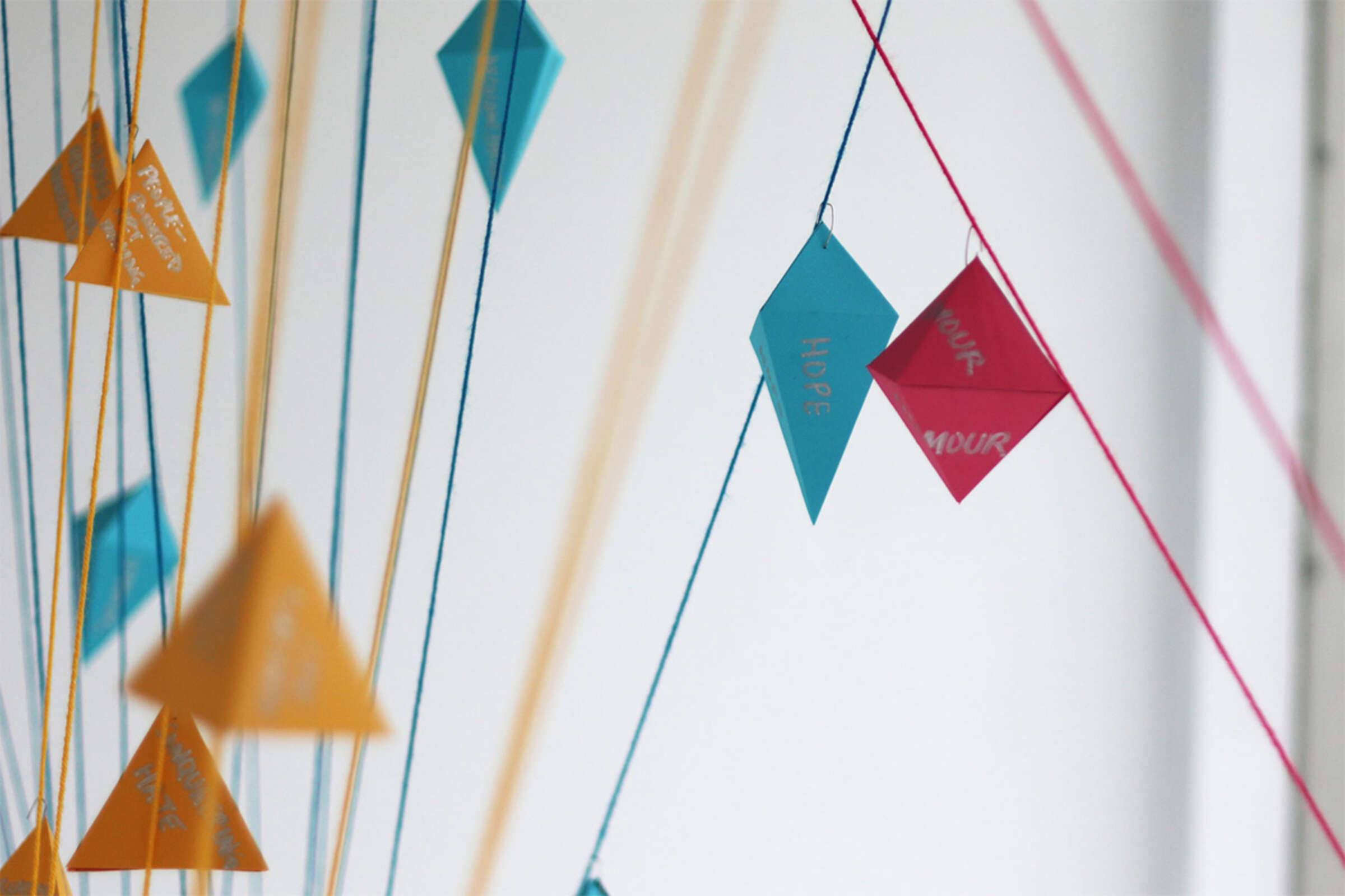 A close-up image of colorful paper shapes hanging on strings against a white background. The shapes include orange triangles, blue diamonds, and red diamonds, some with visible hand-written messages like "HOPE" and "YOUR" in white text.